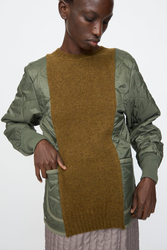 A person wears the Bless No. 78 Liningknitter Sweater in Olive, featuring a green quilted polyester design with an olive-brown knit center panel, standing against a plain white background.