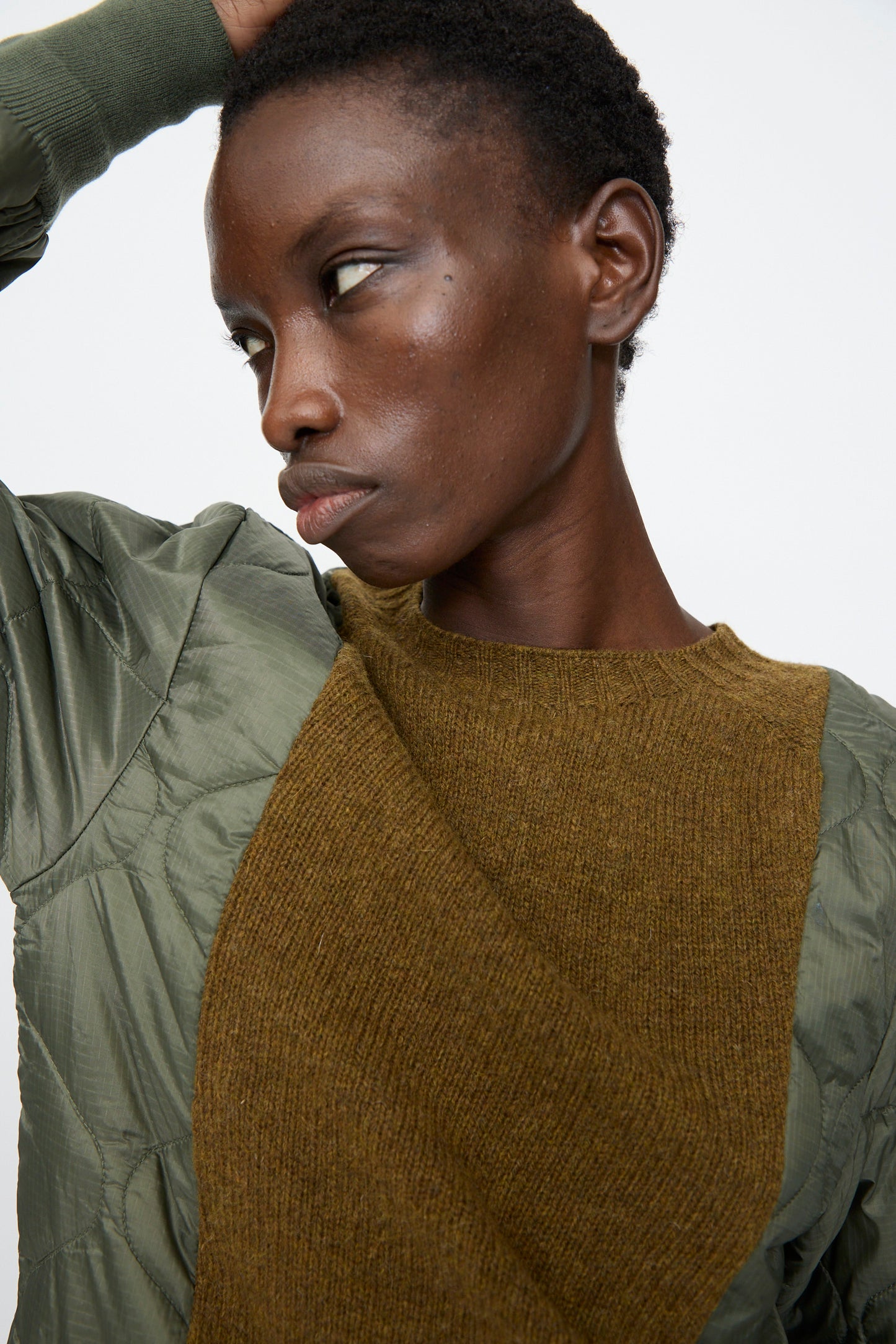 A person wearing the Bless No. 78 Liningknitter Sweater in Olive, featuring quilted polyester and a brown knit panel, looks to the side with one hand on their head against a plain background.