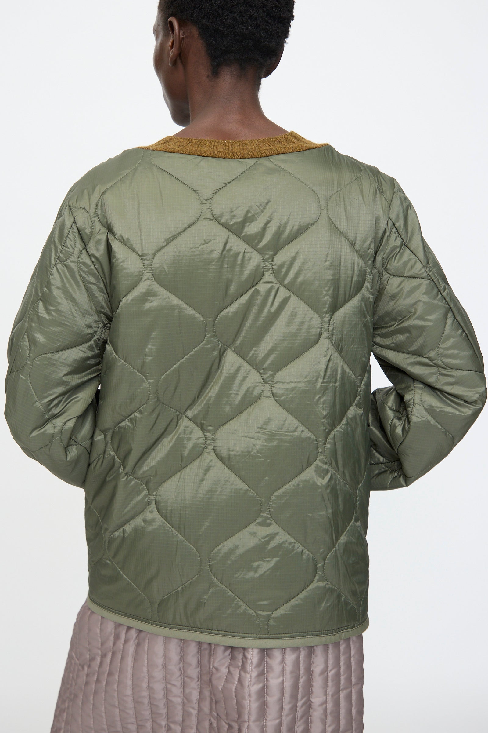 Person seen from the back wearing a quilted olive green jacket with a mustard collar, layered over the Bless No. 78 Liningknitter Sweater in Olive, paired with a light mauve quilted skirt.