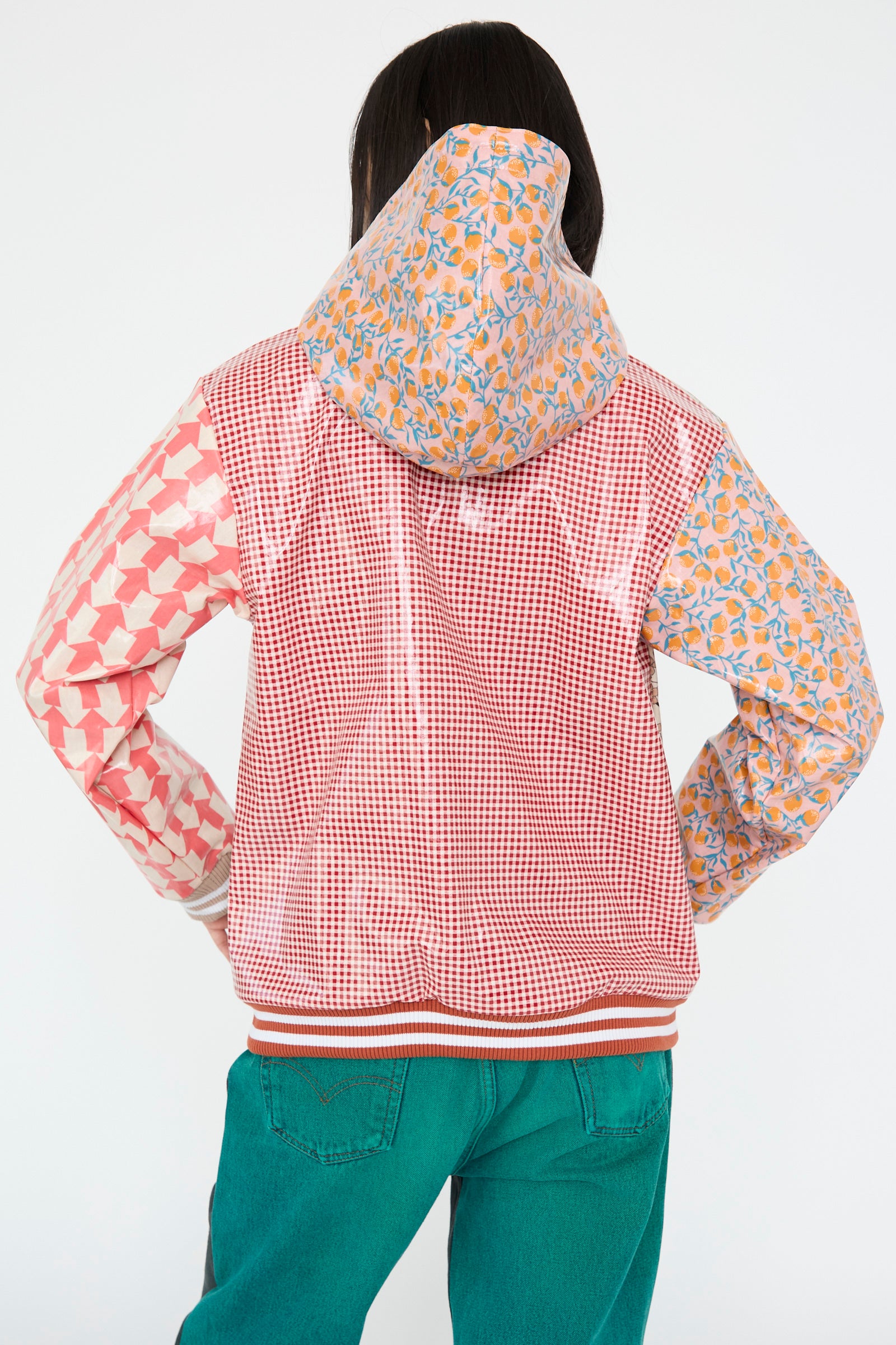 A person facing away wears the No. 79 Cut Rain Jacket Hood in a red color mix from Bless, featuring PVC-coated geometric and floral patterns, paired with teal pants. 