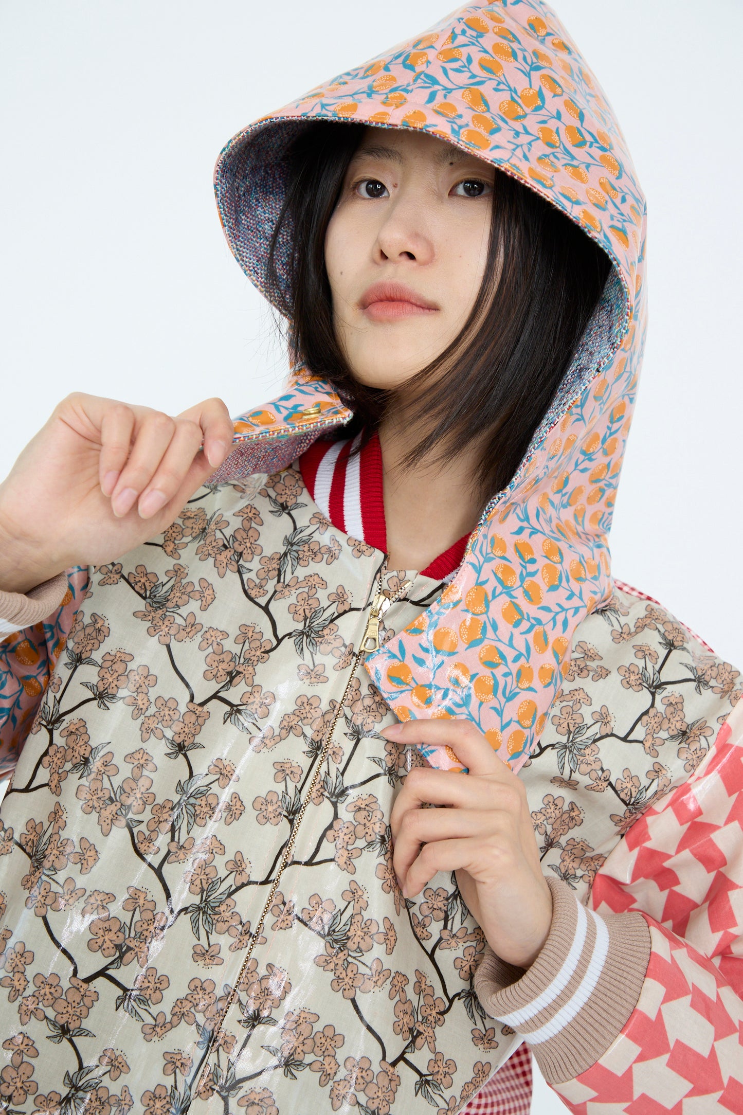 The model wears the Bless No. 79 Cut Rain Jacket Hood in Red Color Mix and looks at the camera while holding the PVC-coated floral hood, against a white backdrop. 