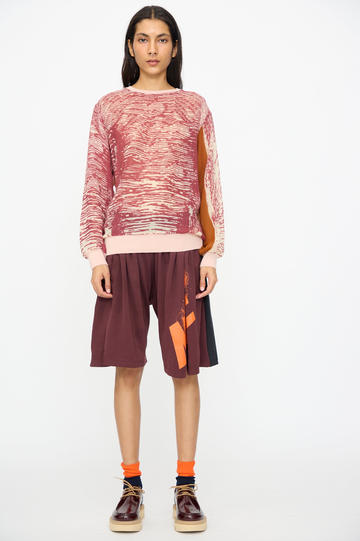 A person stands against a white background wearing the Bless No. 81 M-Sweater in Exelephant, paired with maroon shorts featuring orange accents, orange socks, and burgundy shoes.