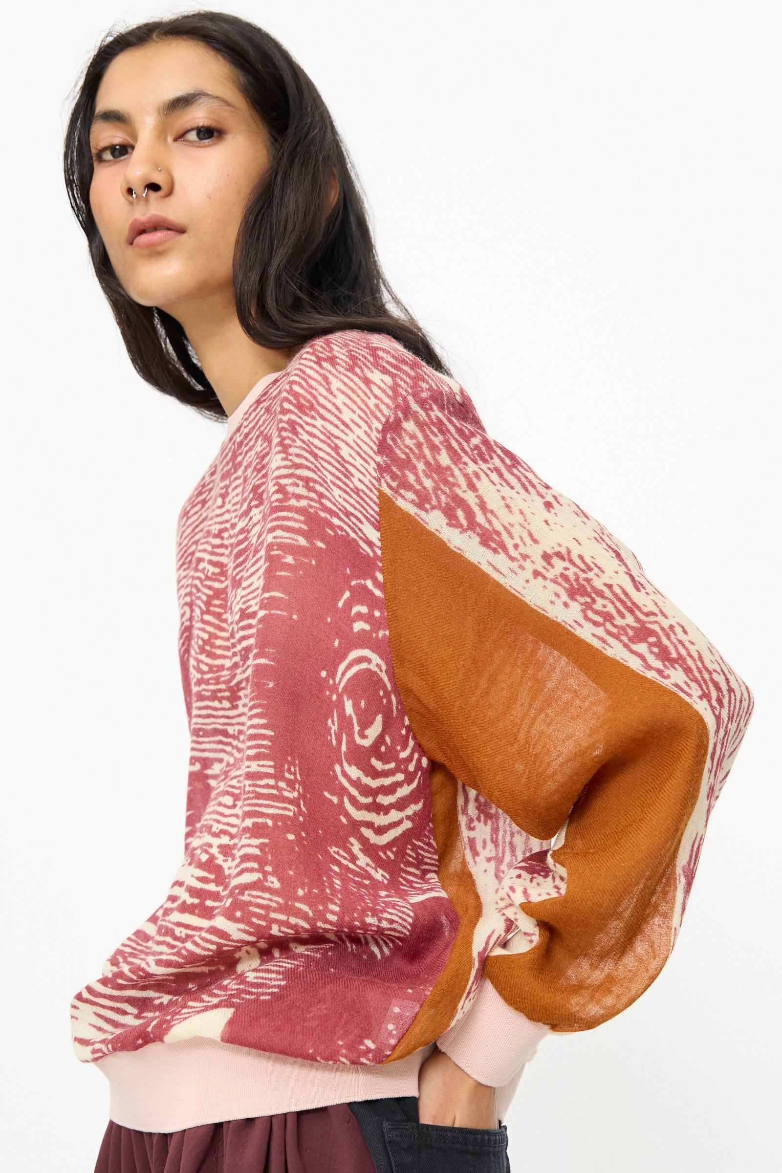 A person with long dark hair and a nose ring wears the Bless No. 81 M-Sweater in Exelephant, featuring a pink, cream, and brown print, while standing sideways against a plain white background.