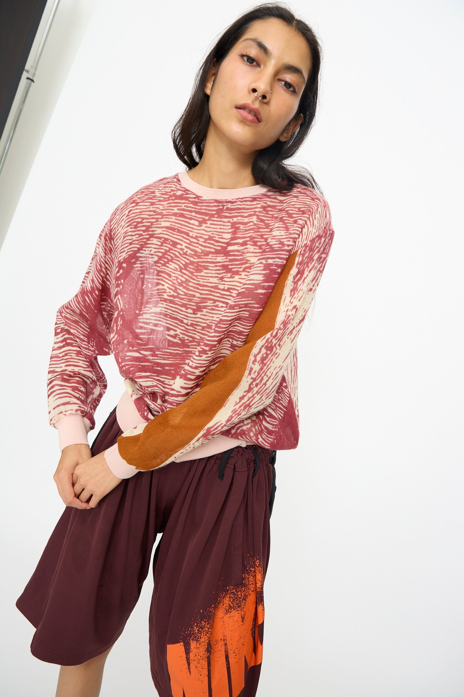 A person wears the Bless No. 81 M-Sweater in Exelephant, featuring a pink elephant skin print with orange details, and burgundy shorts with orange print, standing against a plain white background.