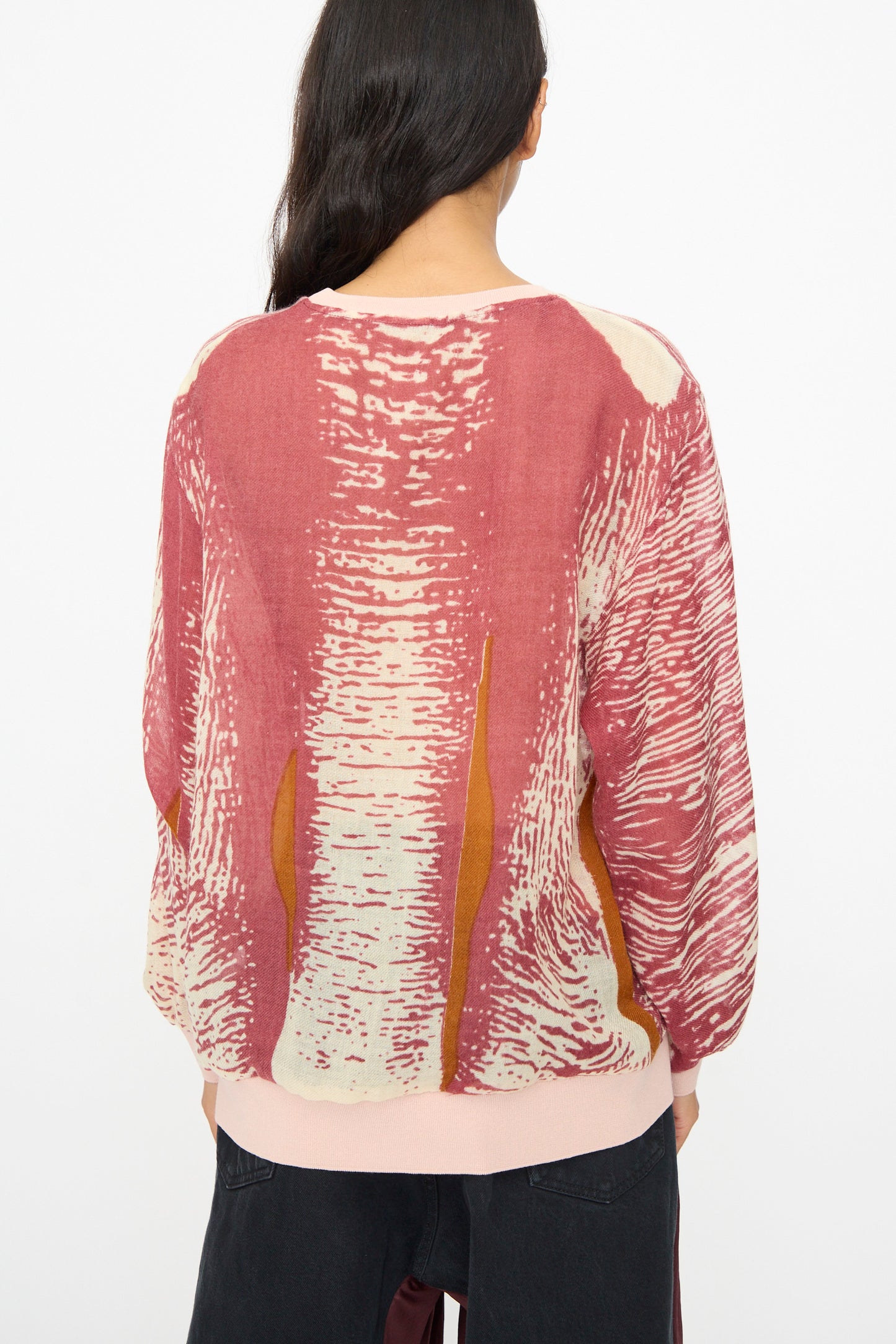 Person with long dark hair wears the Bless No. 81 M-Sweater in Exelephant, a pink and red printed wool sweater, with black pants, photographed from the back against a plain white background.