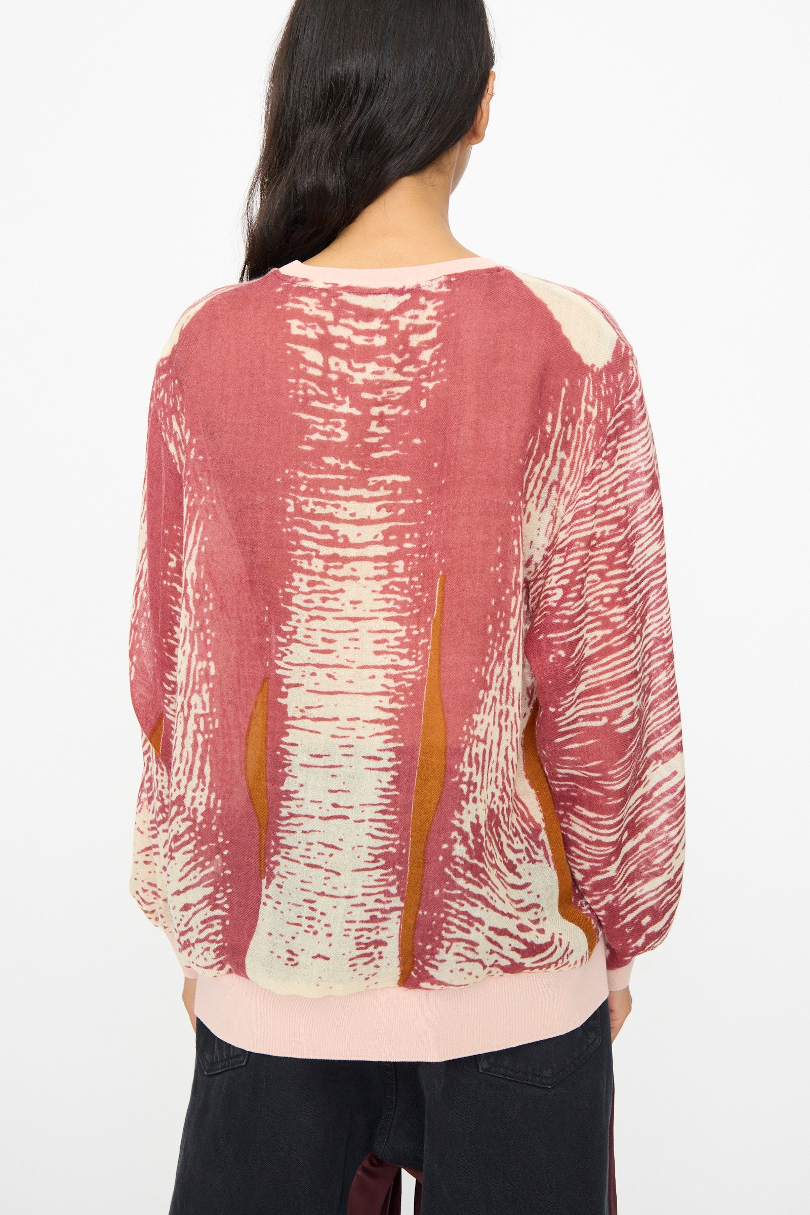 Person with long dark hair wears the Bless No. 81 M-Sweater in Exelephant, a pink and red printed wool sweater, with black pants, photographed from the back against a plain white background.