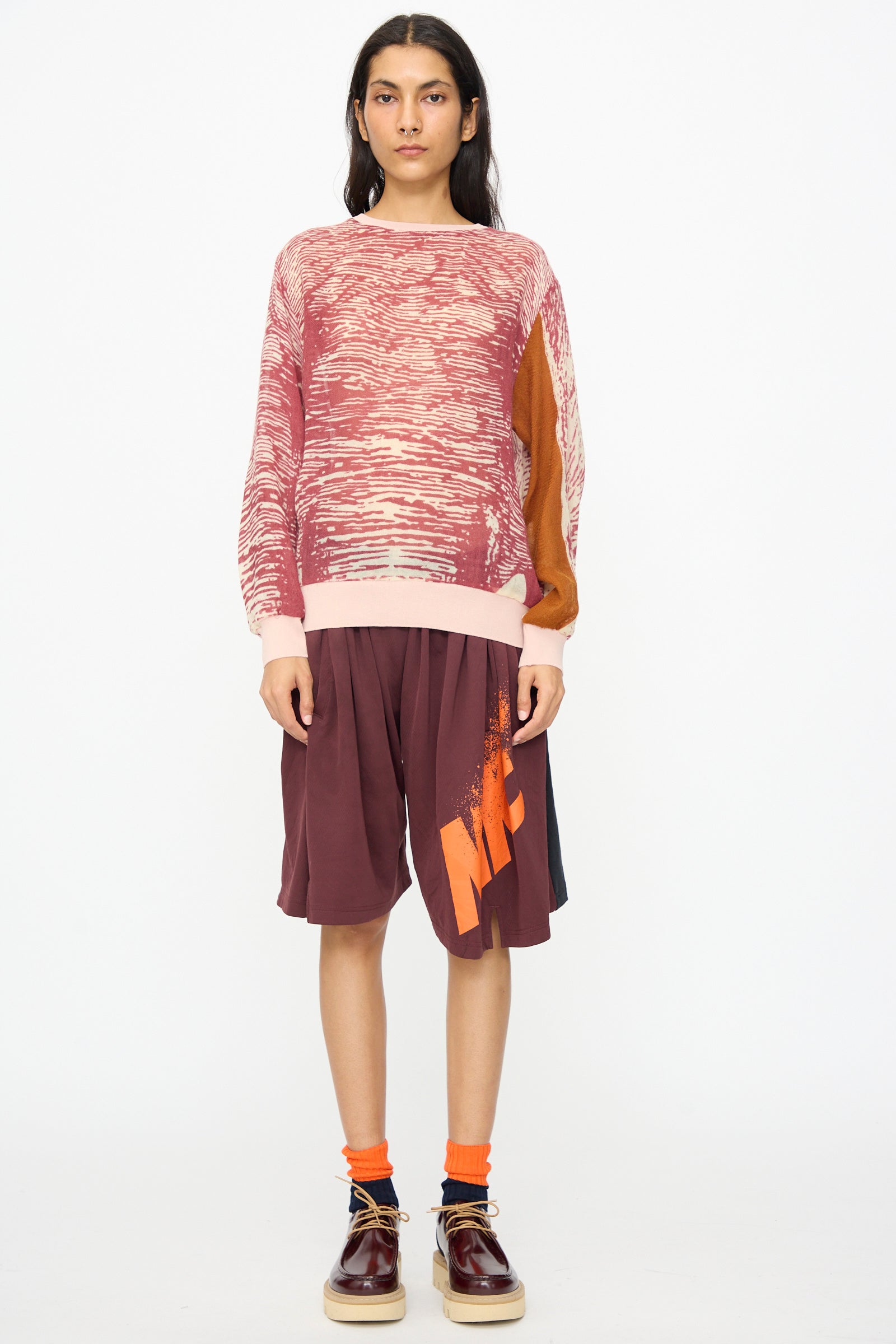 A person stands facing forward in a textured pink and brown sweater, orange socks, dark brown shoes, and Bless No. 81 Overjoggingshorts in Vintage Basketball Unique I against a plain white background.