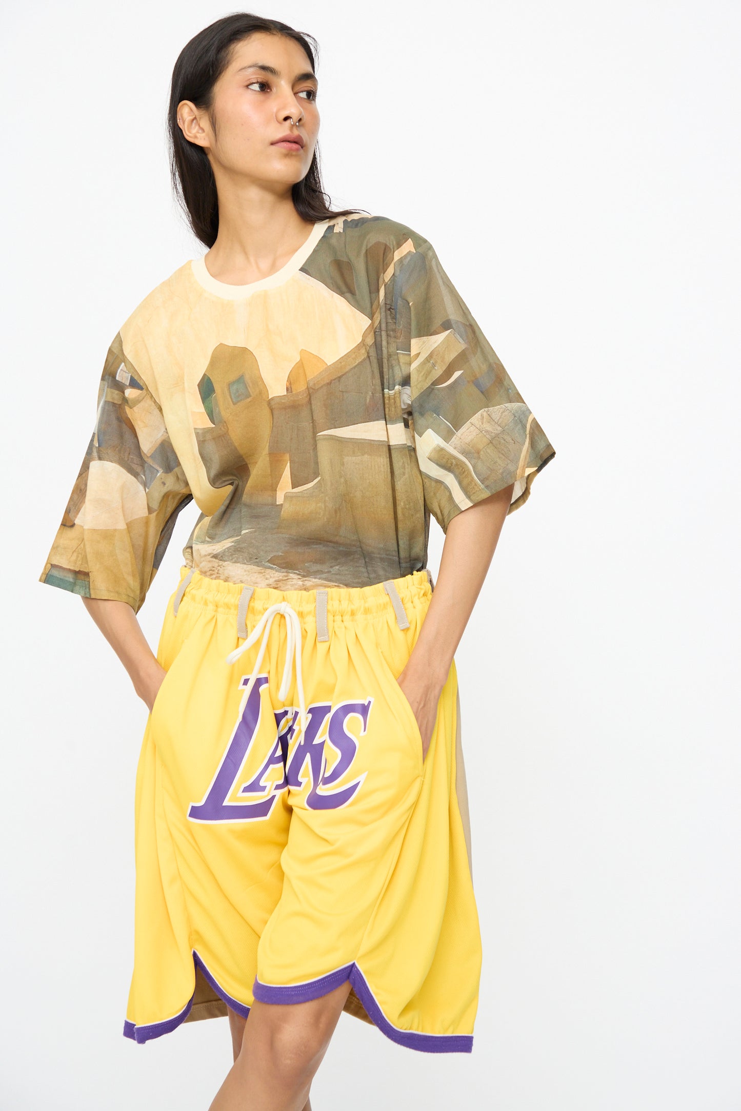 A person stands against a plain background wearing a camouflage t-shirt and Bless No. 81 Overjoggingshorts in Vintage Basketball Unique II, with hands in pockets.