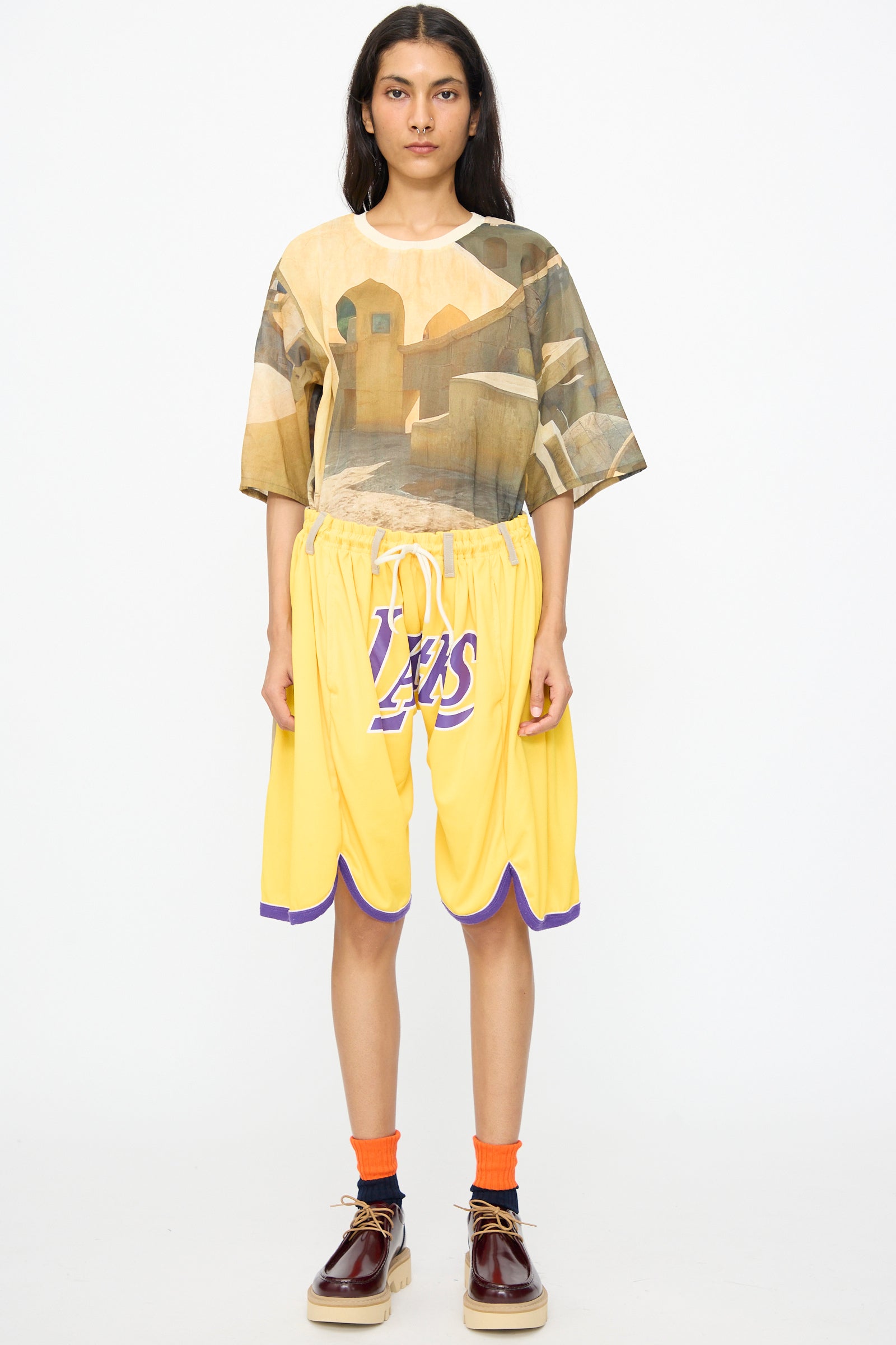 A person stands facing forward in a printed oversized shirt, Bless No. 81 Overjoggingshorts in Vintage Basketball Unique II, orange socks, and brown platform shoes against a plain white background.