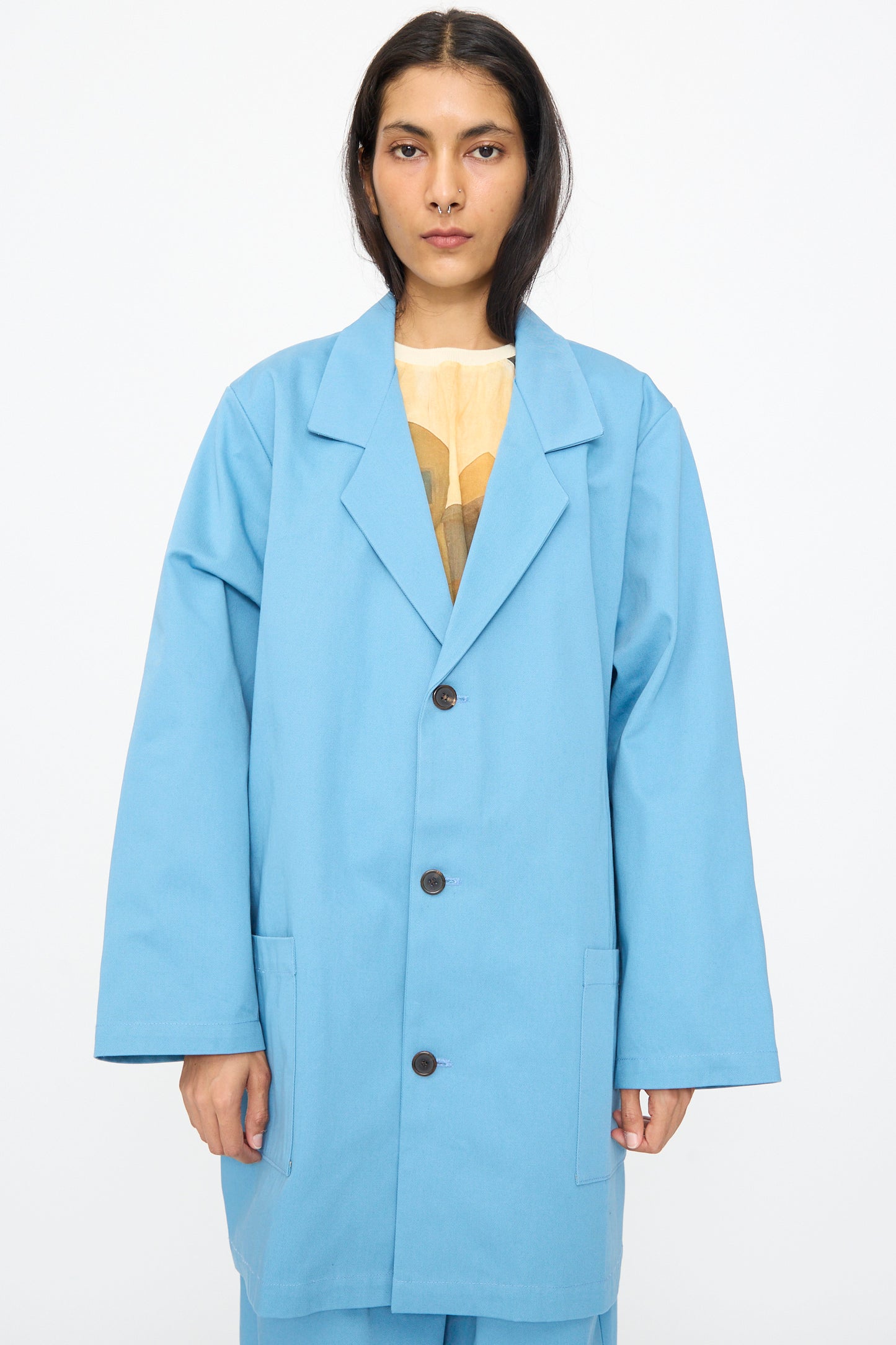 A person stands against a white background wearing the Bless No. 81 Summer Workcoat Short in Skyblue, a loose-fitting cotton coat featuring oversized styling, large lapels, and three buttons, with their hands at their sides.