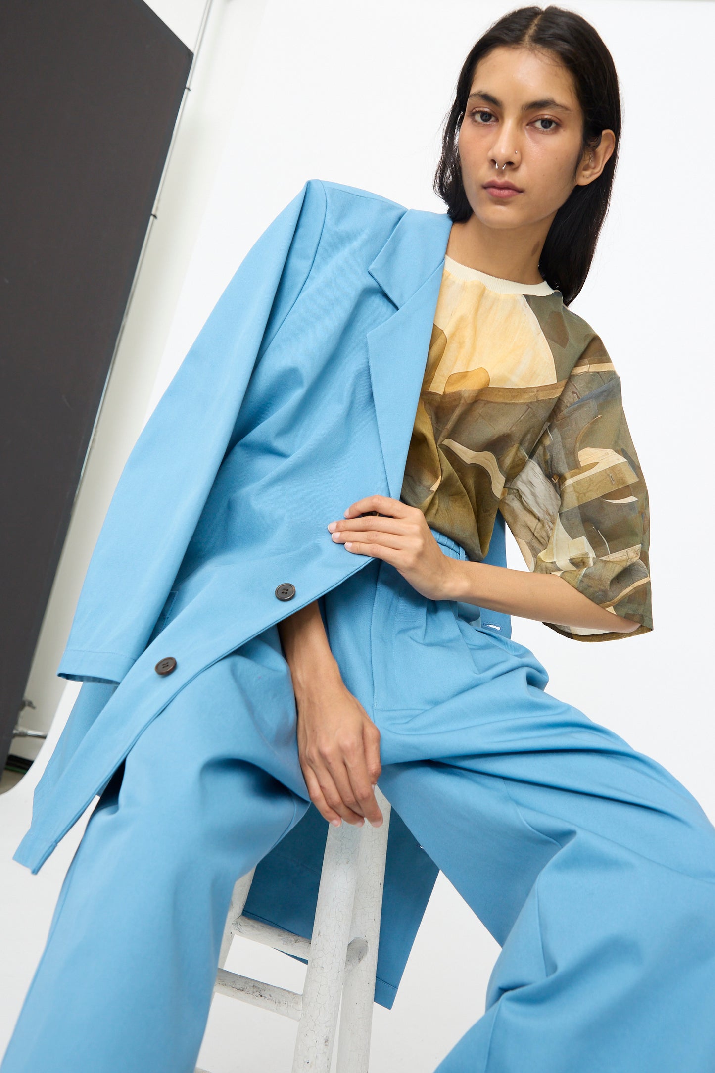 A person wearing the Bless No. 81 Summer Workcoat Short in Skyblue and a patterned beige shirt sits on a stool against a plain background.