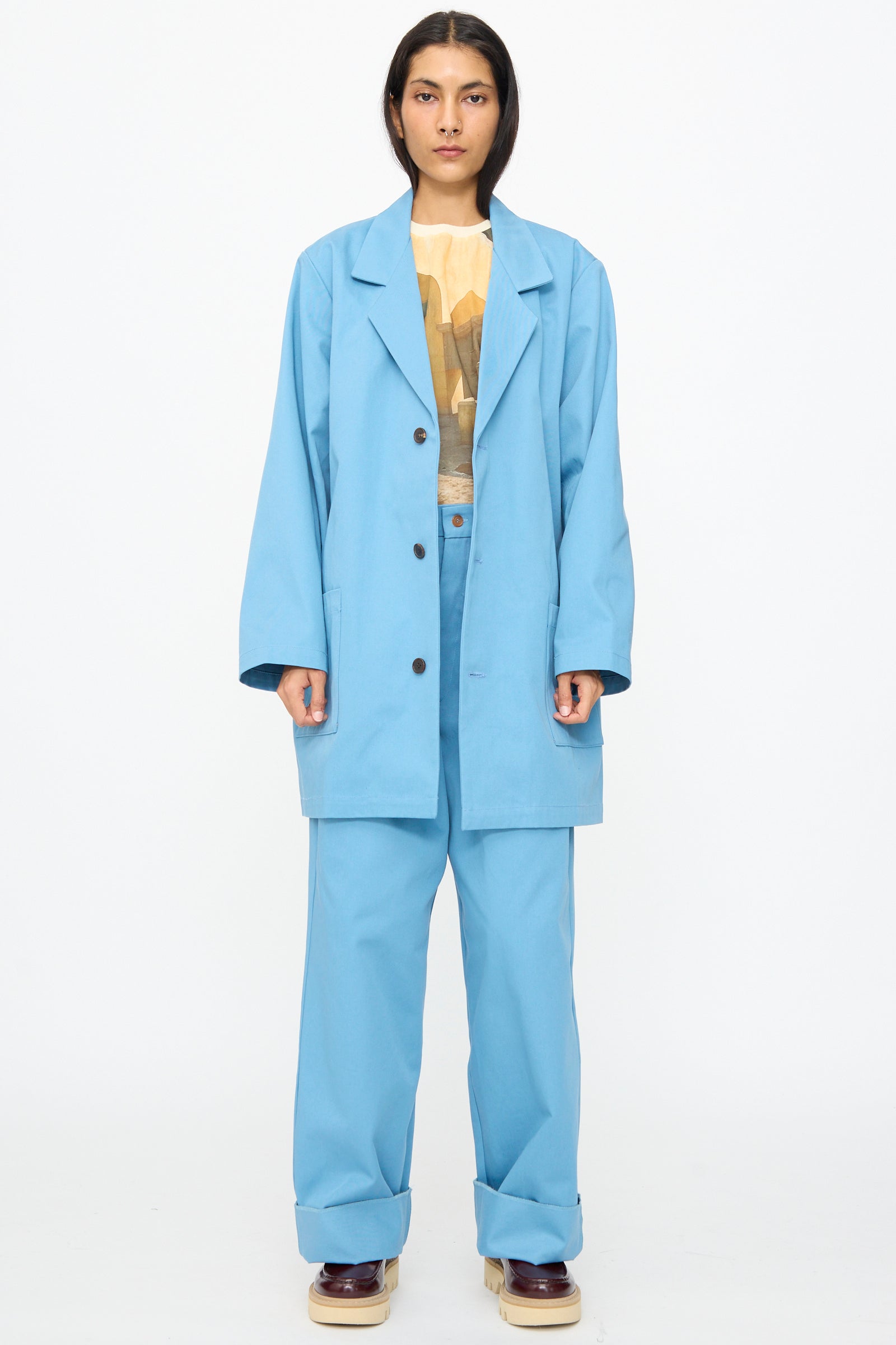 A person stands against a white background wearing Bless’s No. 81 Summer Workcoat Short in Skyblue with matching pants, a printed shirt underneath, and beige platform shoes.