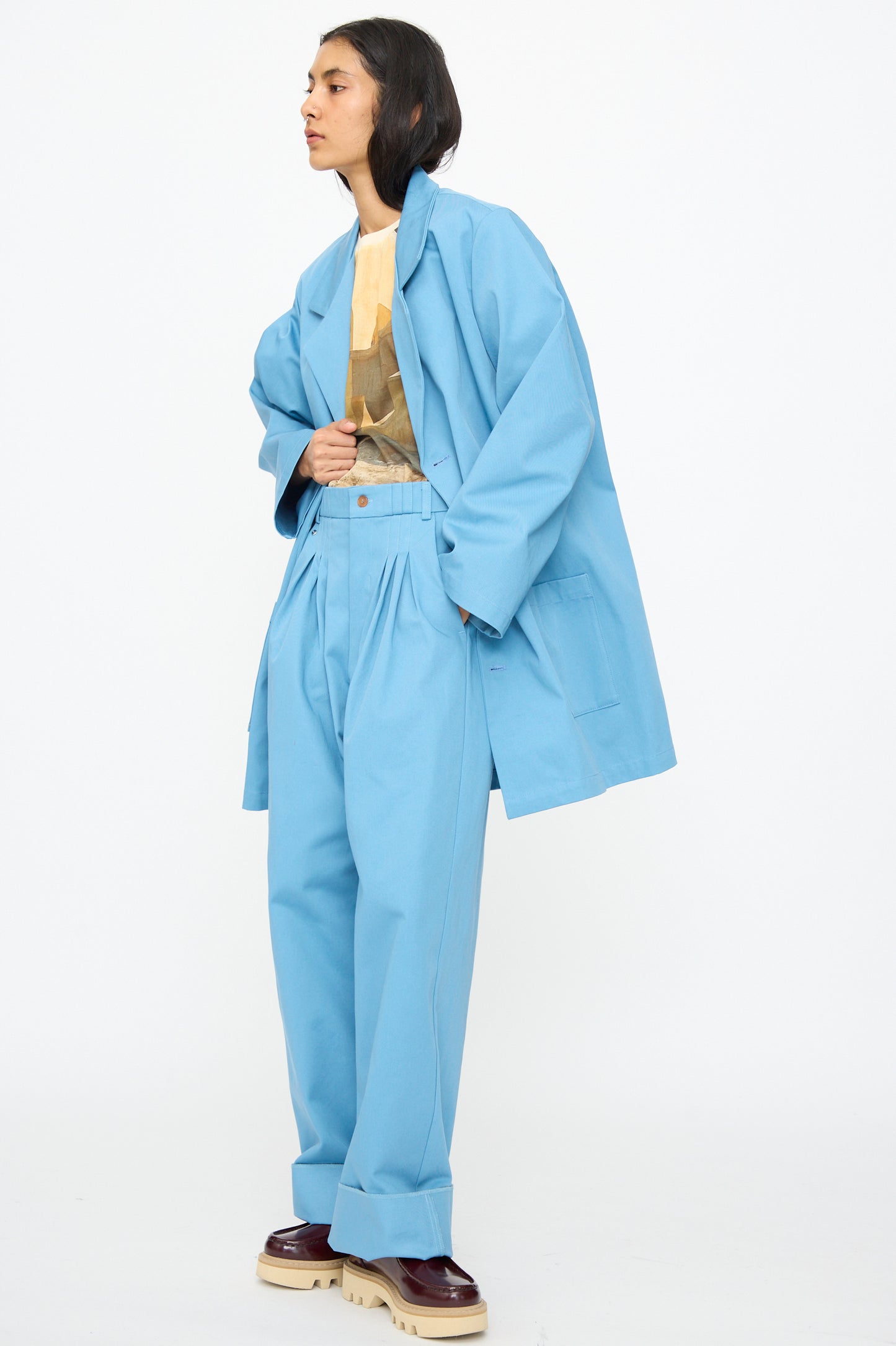 A person wearing Bless's No. 81 Summer Workcoat Short in Skyblue, paired with wide-leg pants, a graphic t-shirt, and platform shoes, stands against a plain white background.