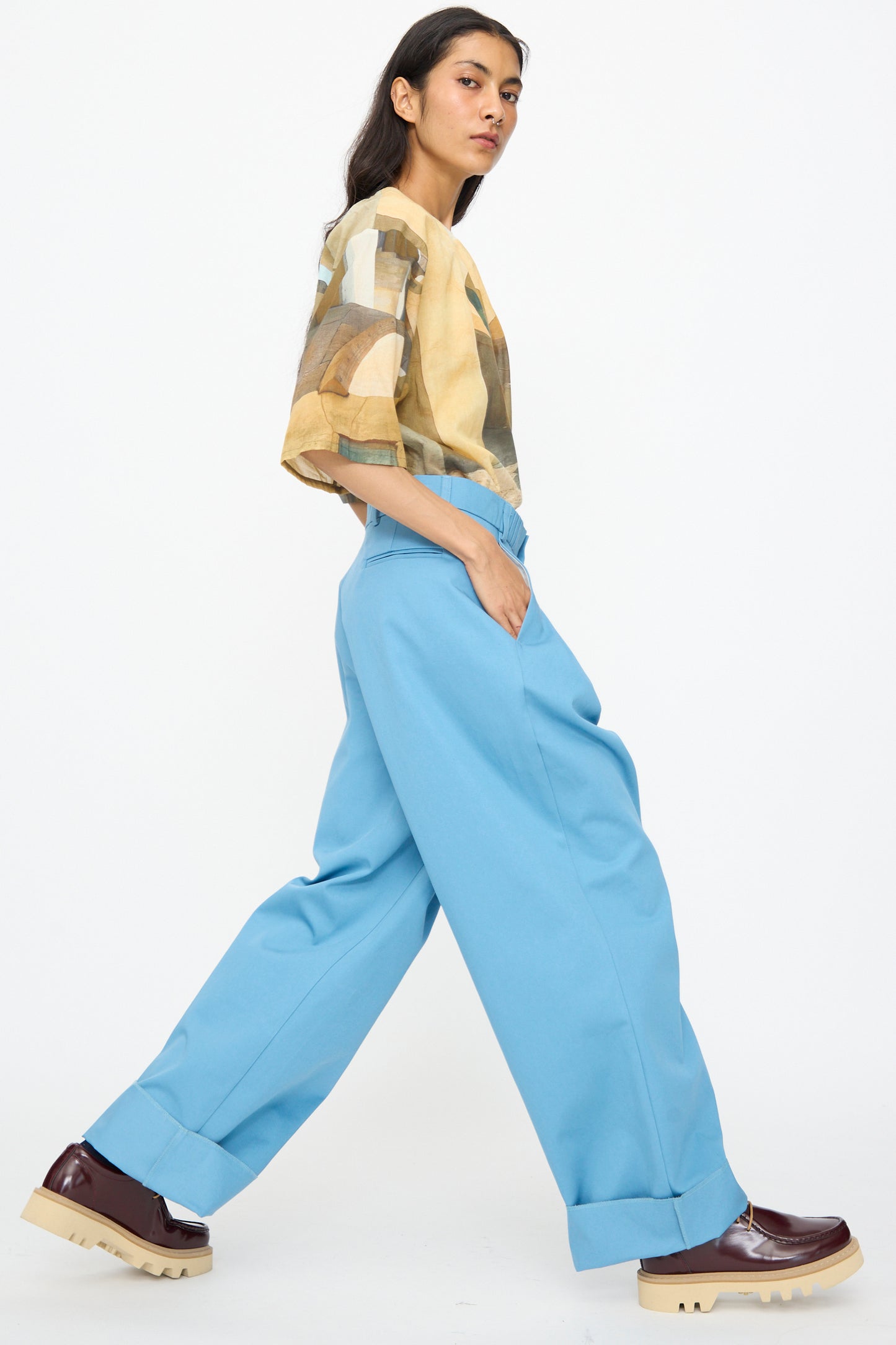 Wearing the Bless No. 81 Ultrawidepleated Pant in Skyblue with a patterned beige shirt and burgundy platform shoes, a person walks sideways against a plain white background.