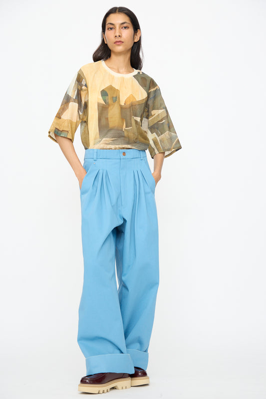 A person stands against a white background wearing a geometric print shirt and Bless's No. 81 Ultrawidepleated Pant in Skyblue, paired with tan platform shoes.