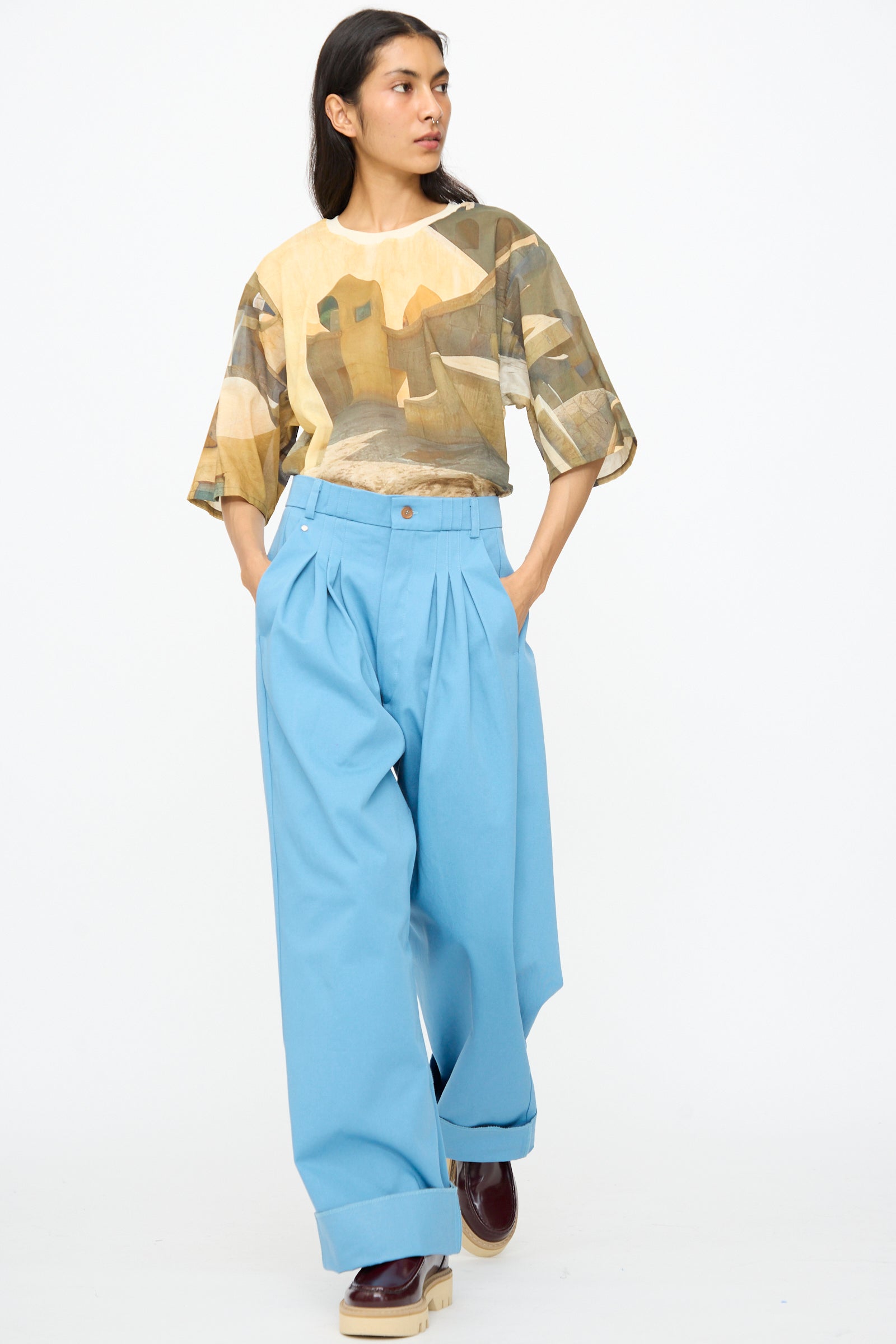 A person stands with hands in pockets against a plain white background, wearing wide-leg Bless No. 81 Ultrawidepleated Pant in Skyblue, a loose printed shirt, and dark brown shoes.