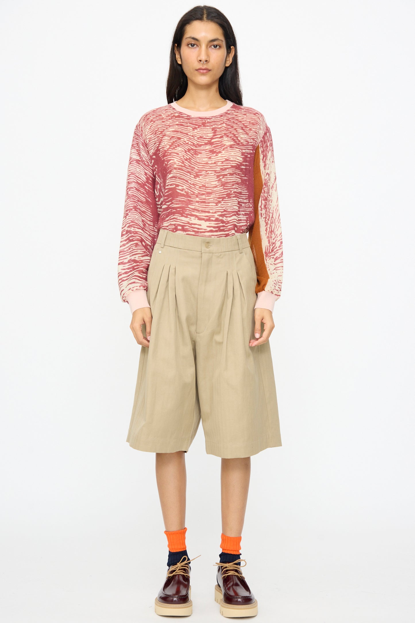 A person stands facing forward wearing a red and beige patterned long-sleeve top, Bless No. 81 Ultrawidepleated Shorts in Sandkoeper, orange socks, and burgundy shoes.