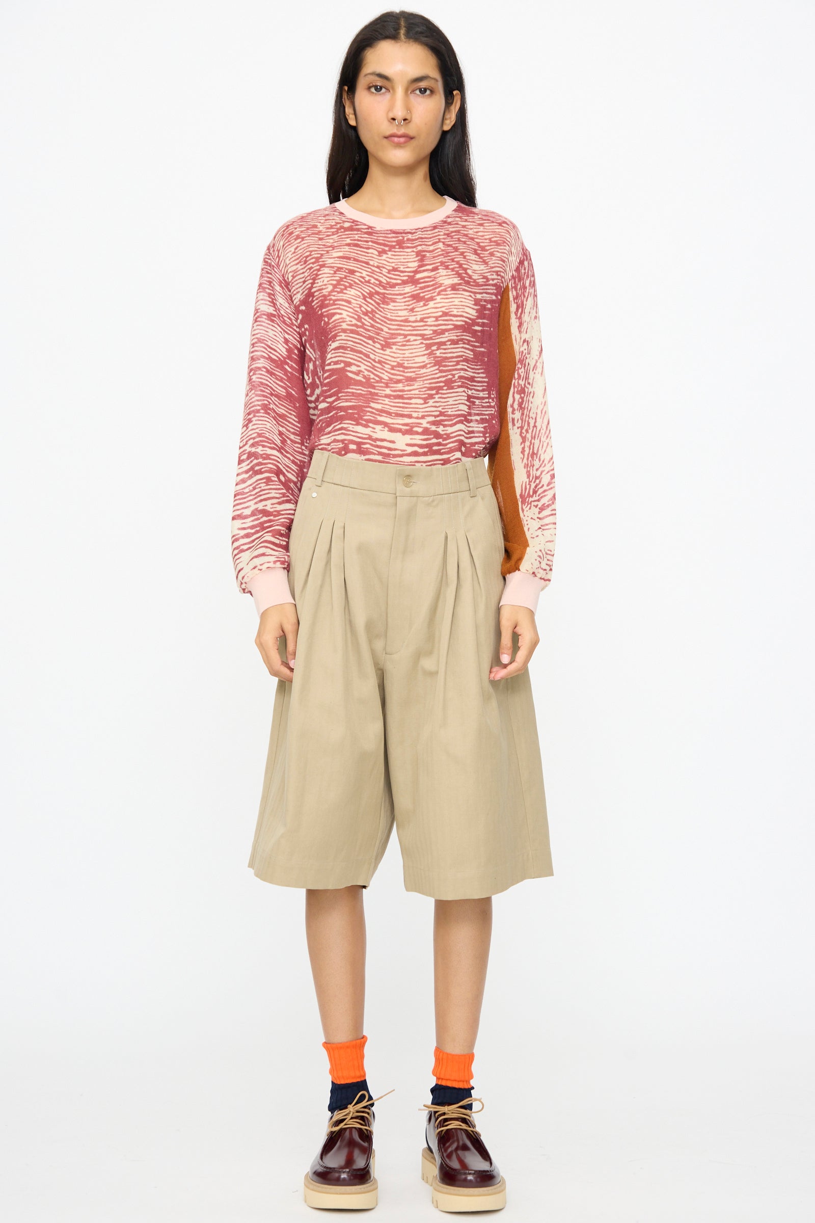 A person stands facing forward wearing a red and beige patterned long-sleeve top, Bless No. 81 Ultrawidepleated Shorts in Sandkoeper, orange socks, and burgundy shoes.