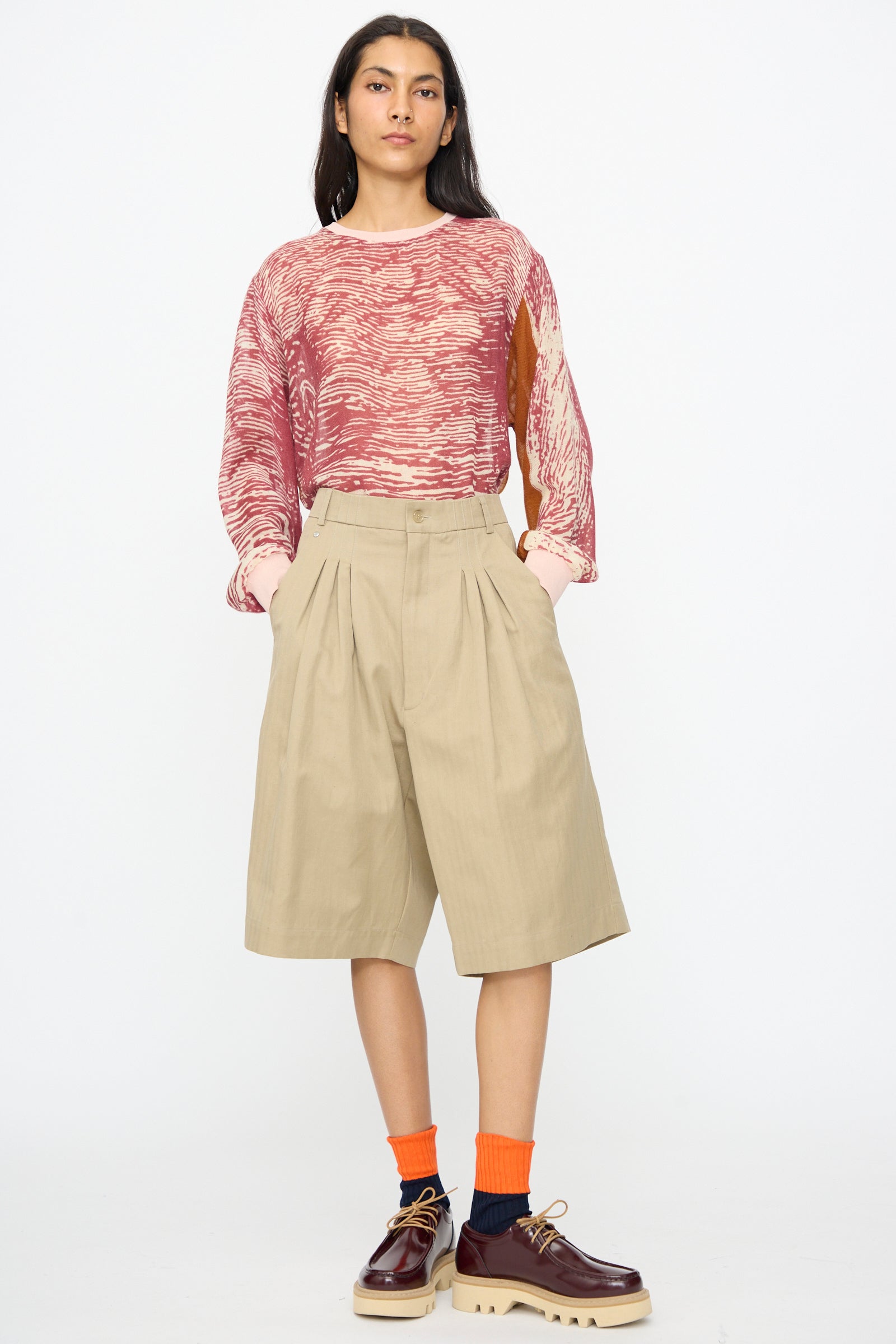 A person stands facing forward, wearing a red patterned long-sleeve shirt, Bless No. 81 Ultrawidepleated Shorts in Sandkoeper, orange socks, and brown platform shoes against a plain background.