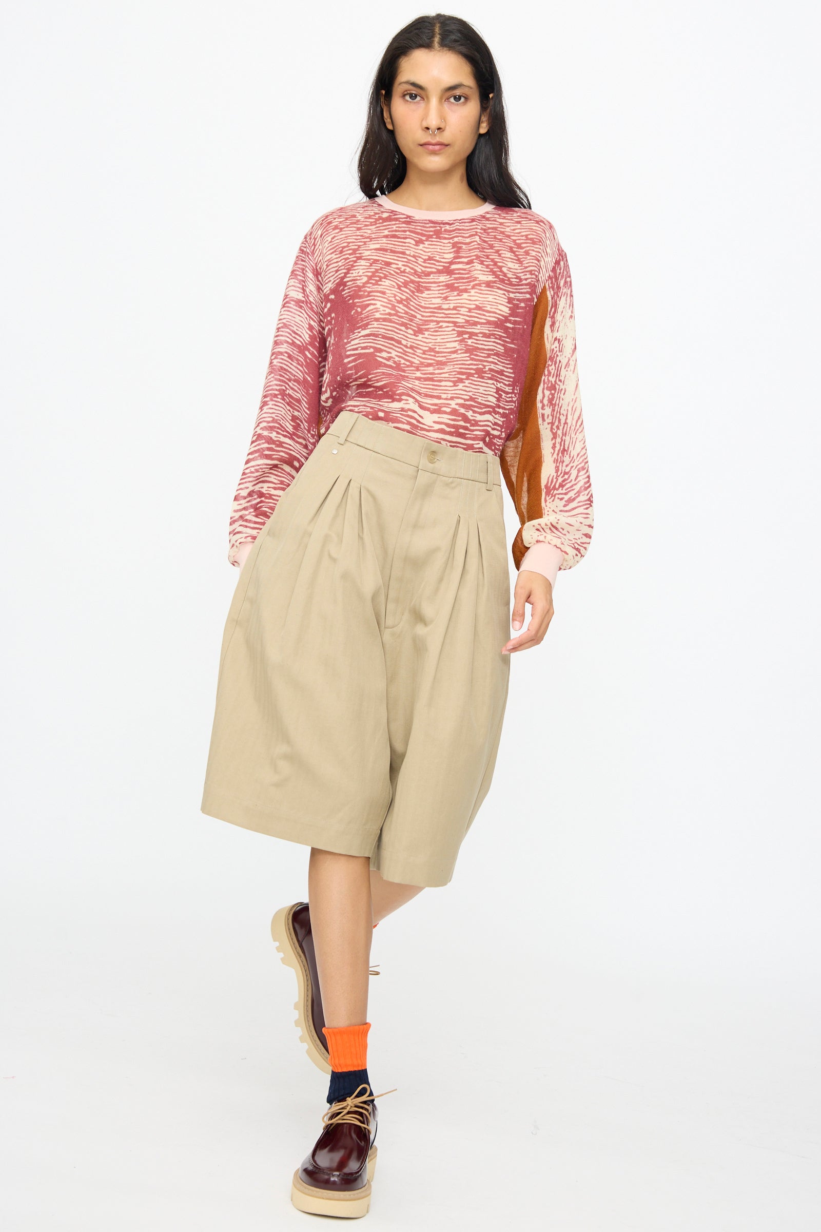 A woman stands against a plain white background wearing a red patterned long-sleeve shirt, orange socks, brown shoes, and Bless No. 81 Ultrawidepleated Shorts in Sandkoeper.
