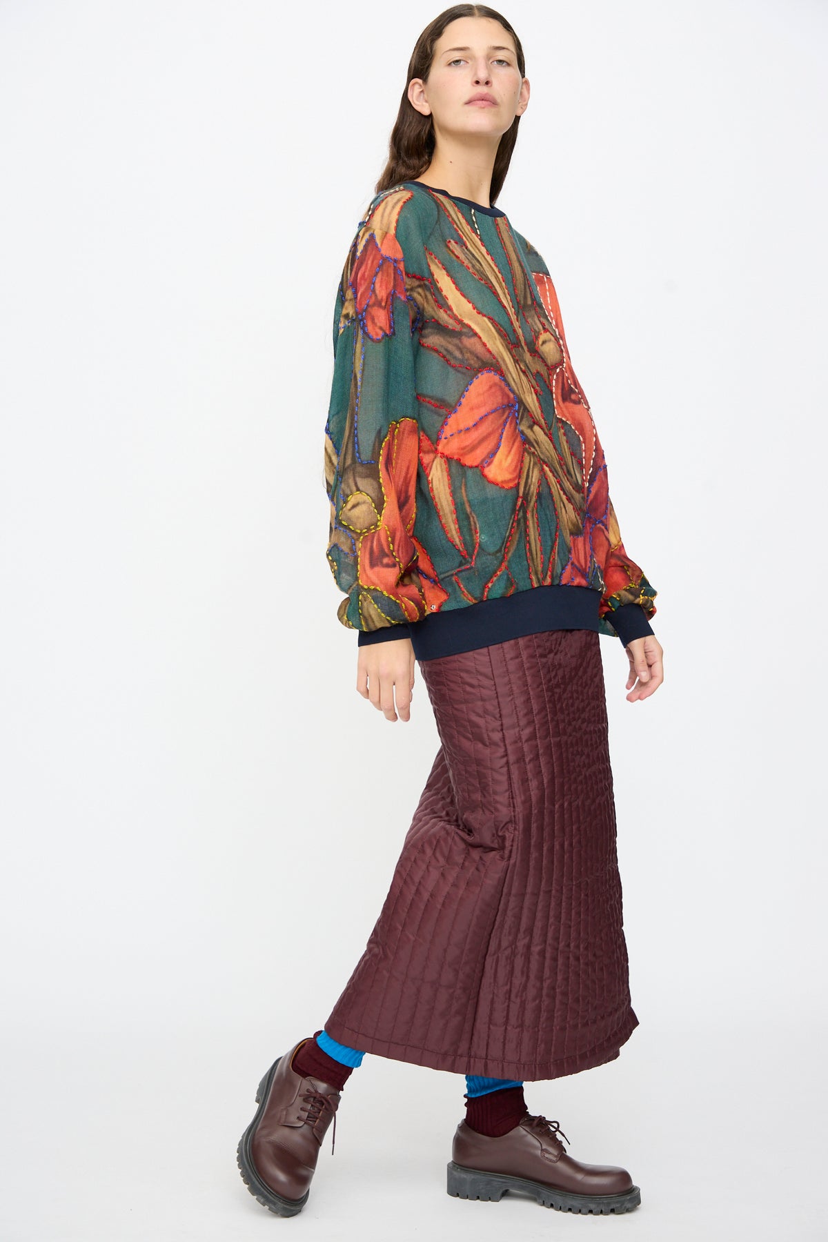A person wearing a Bless No. 82 Embroidered Sweater in Dark Brown and Green with a floral print, a long quilted dark brown skirt, and dark brown lace-up shoes.