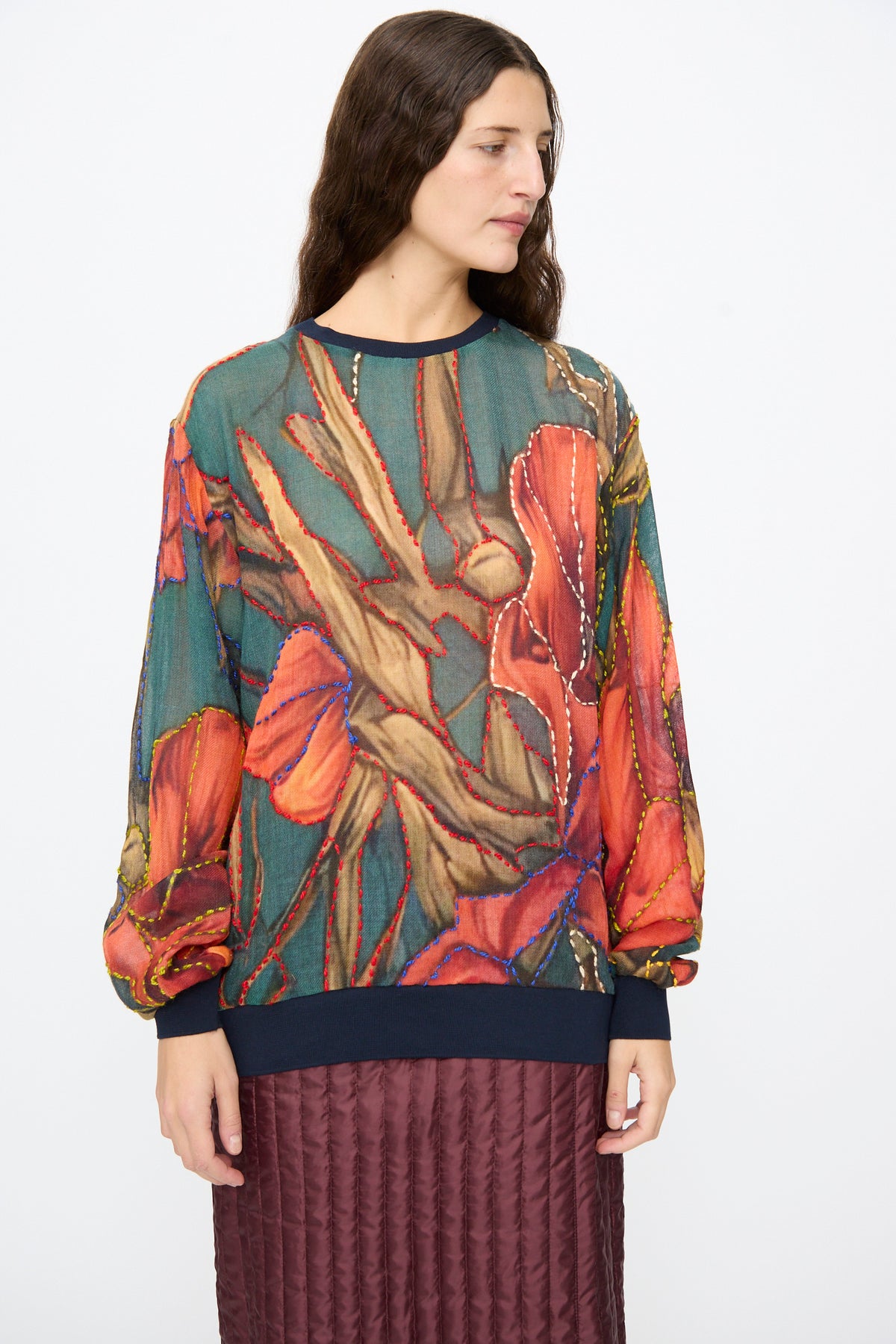 A person wearing a Bless No. 82 Embroidered Sweater in Dark Brown and Green with a botanical motif and contrast-colored embroidery stitches, paired with a dark quilted skirt.