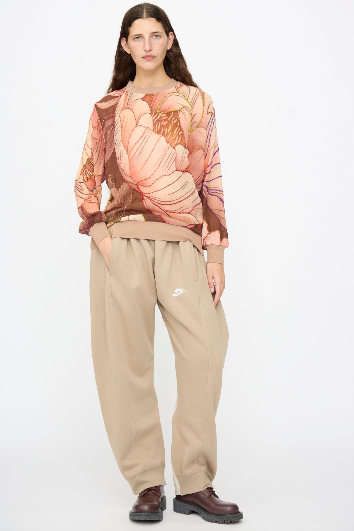A person wearing a Bless No. 82 Embroidered Sweater in Multicolor Beige and Rosé with a large pink and brown pattern, paired with tan sweatpants and dark brown lug-sole shoes.