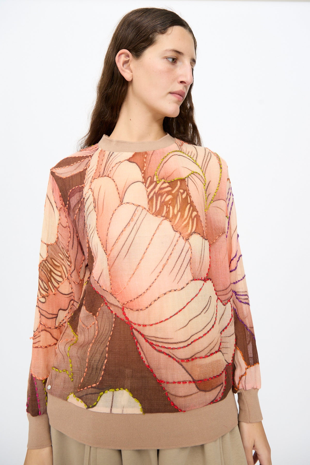 A woman wears the Bless No. 82 Embroidered Sweater in Multicolor Beige and Rosé, a lightweight, long-sleeved piece with a large, abstract floral print outlined in multicolored embroidery.