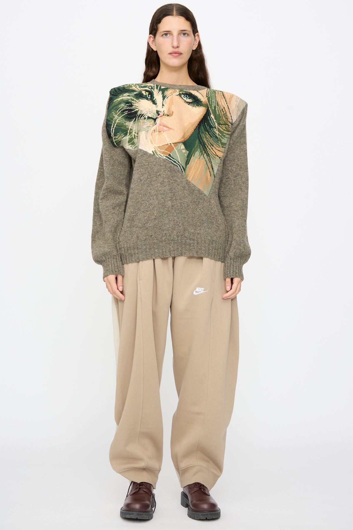 A person wears a Bless No. 82 Kitschenrich in Unique Ultralarge Patchwork I wool pullover featuring a needlepoint cat face and eye, paired with loose tan sweatpants and dark brown shoes.