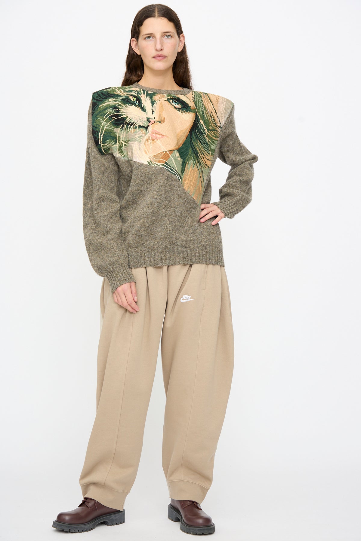 A person wears a Bless No. 82 Kitschenrich in Unique Ultralarge Patchwork I textured brown wool pullover featuring an asymmetrical face and cat graphic, paired with tan sweatpants and dark brown shoes.