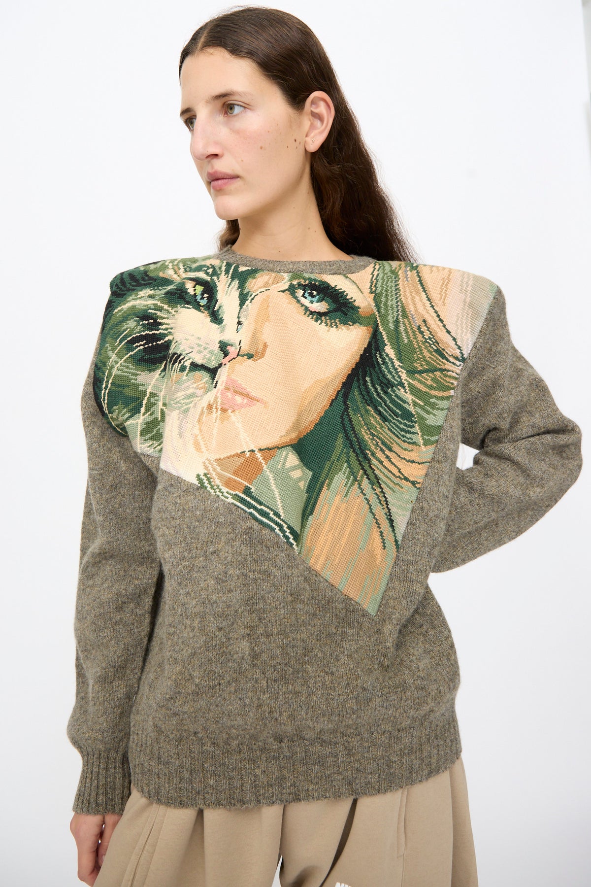 A person wearing a grey wool Bless No. 82 Kitschenrich in Unique Ultralarge Patchwork I pullover sweater featuring a large, triangular needlepoint motif of a woman’s face on the chest.