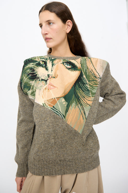 A person wearing a grey wool Bless No. 82 Kitschenrich in Unique Ultralarge Patchwork I pullover sweater featuring a large, triangular needlepoint motif of a woman’s face on the chest.
