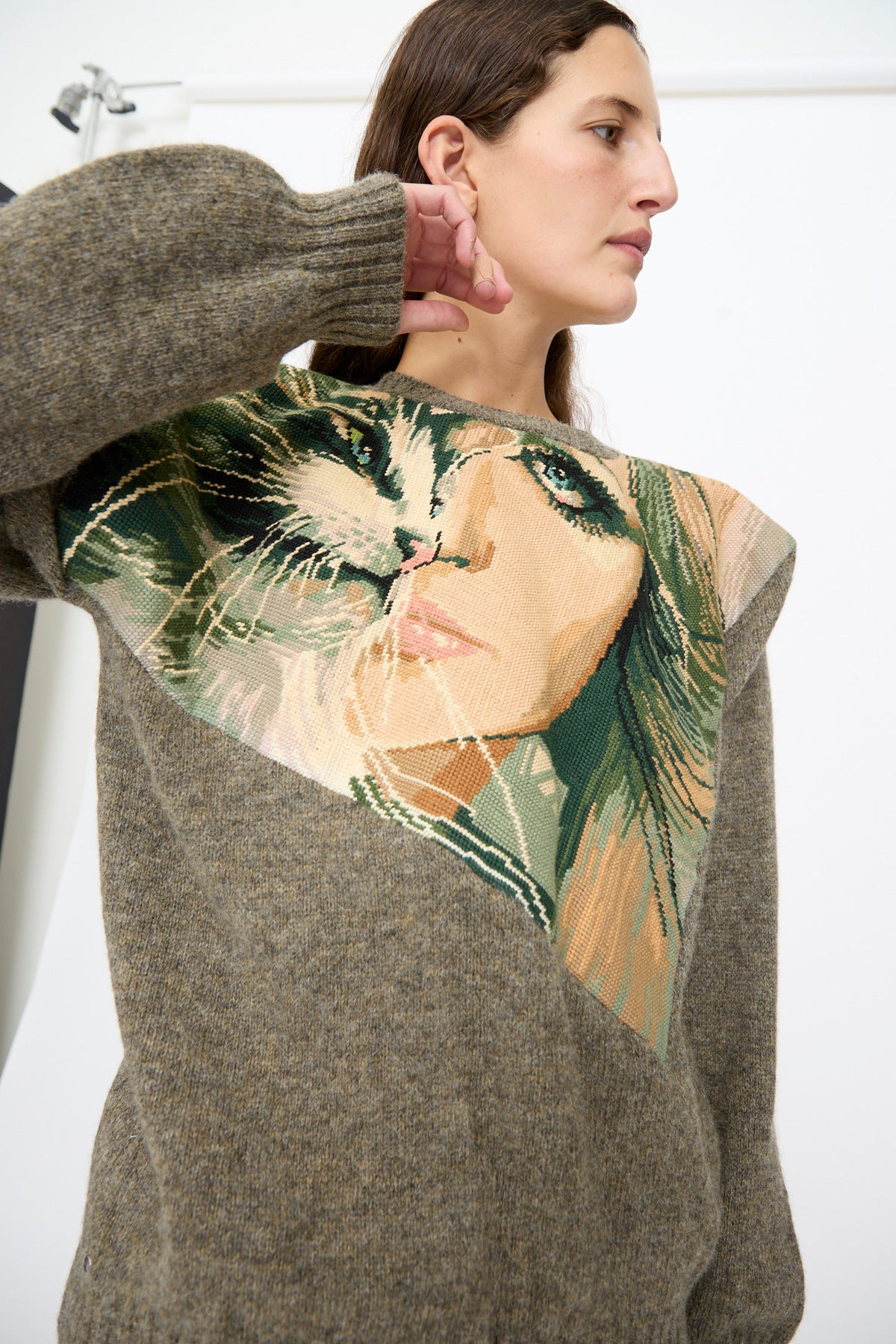 A person wearing the Bless No. 82 Kitschenrich in Unique Ultralarge Patchwork I, a brown wool pullover sweater with a large triangular needlepoint motif of a cat and a woman’s face on the chest.