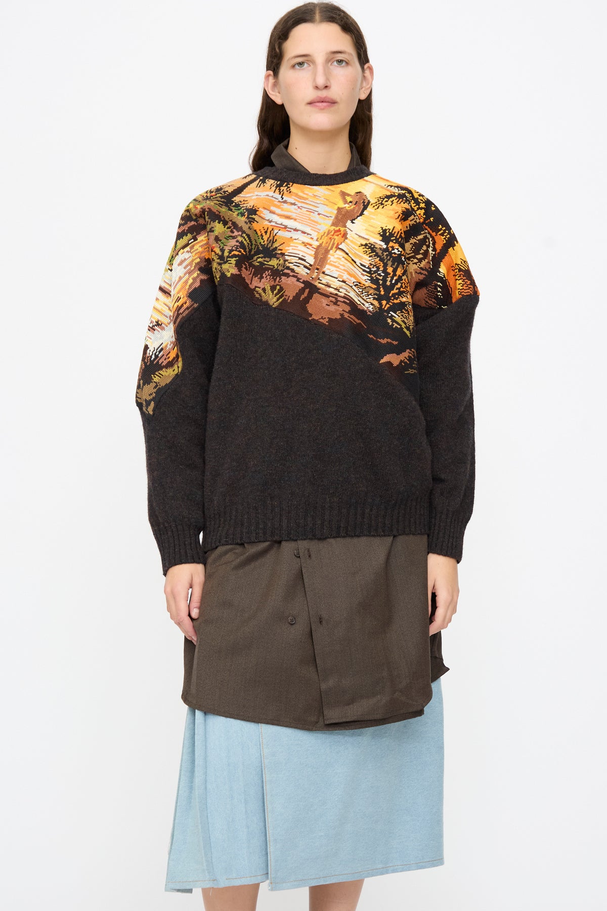 Bless No. 82 Kitschenrich in Unique Ultralarge Patchwork II wool pullover with an orange and brown tropical landscape motif, layered over a dark brown button-up shirt and light blue denim skirt.