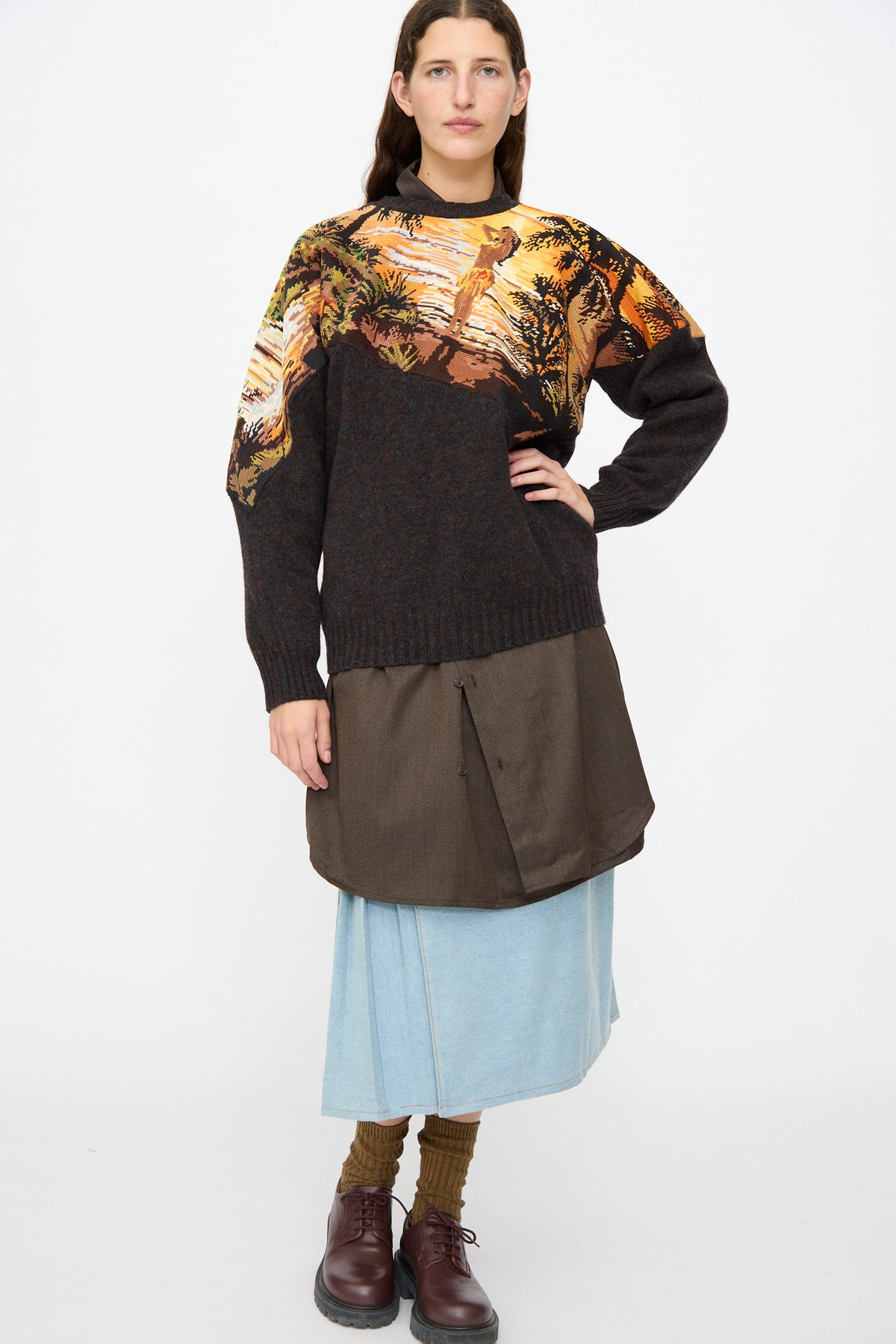 A person wearing a Bless No. 82 Kitschenrich in Unique Ultralarge Patchwork II wool pullover, brown button-up shirt, light blue midi skirt, brown socks, and dark brown shoes.