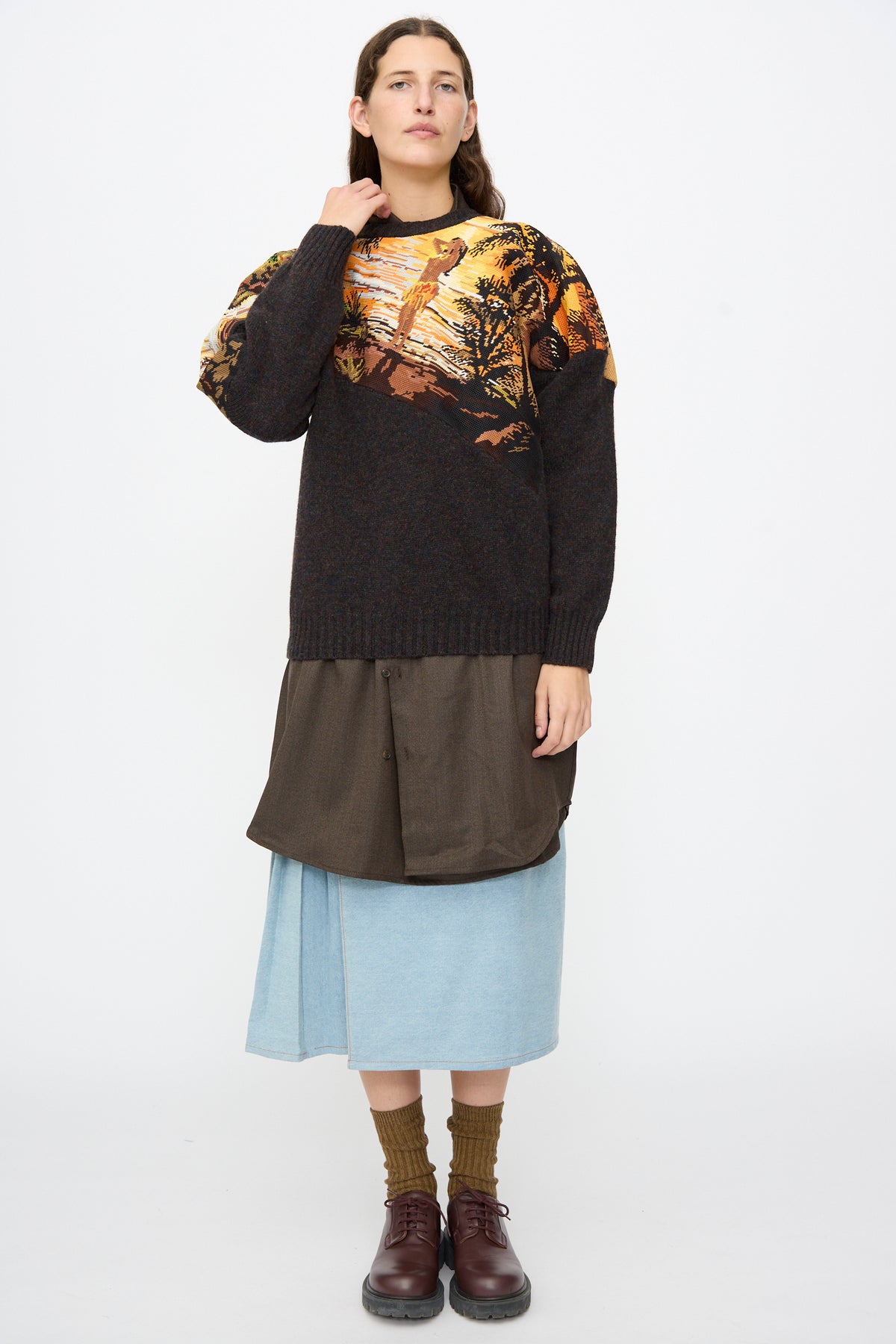 A person wearing a Bless No. 82 Kitschenrich in Unique Ultralarge Patchwork II wool pullover over a brown shirt, a blue denim skirt, brown socks, and dark brown shoes.