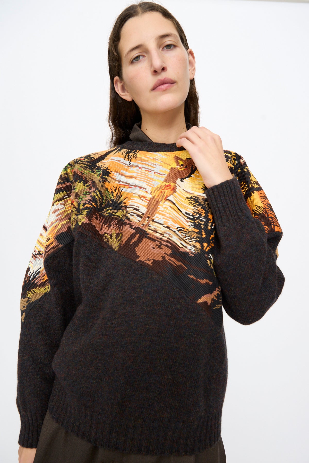 A person wears a Bless No. 82 Kitschenrich in Unique Ultralarge Patchwork II wool pullover with a dark charcoal bottom and an upper panel featuring a vibrant orange, yellow, and brown landscape motif.