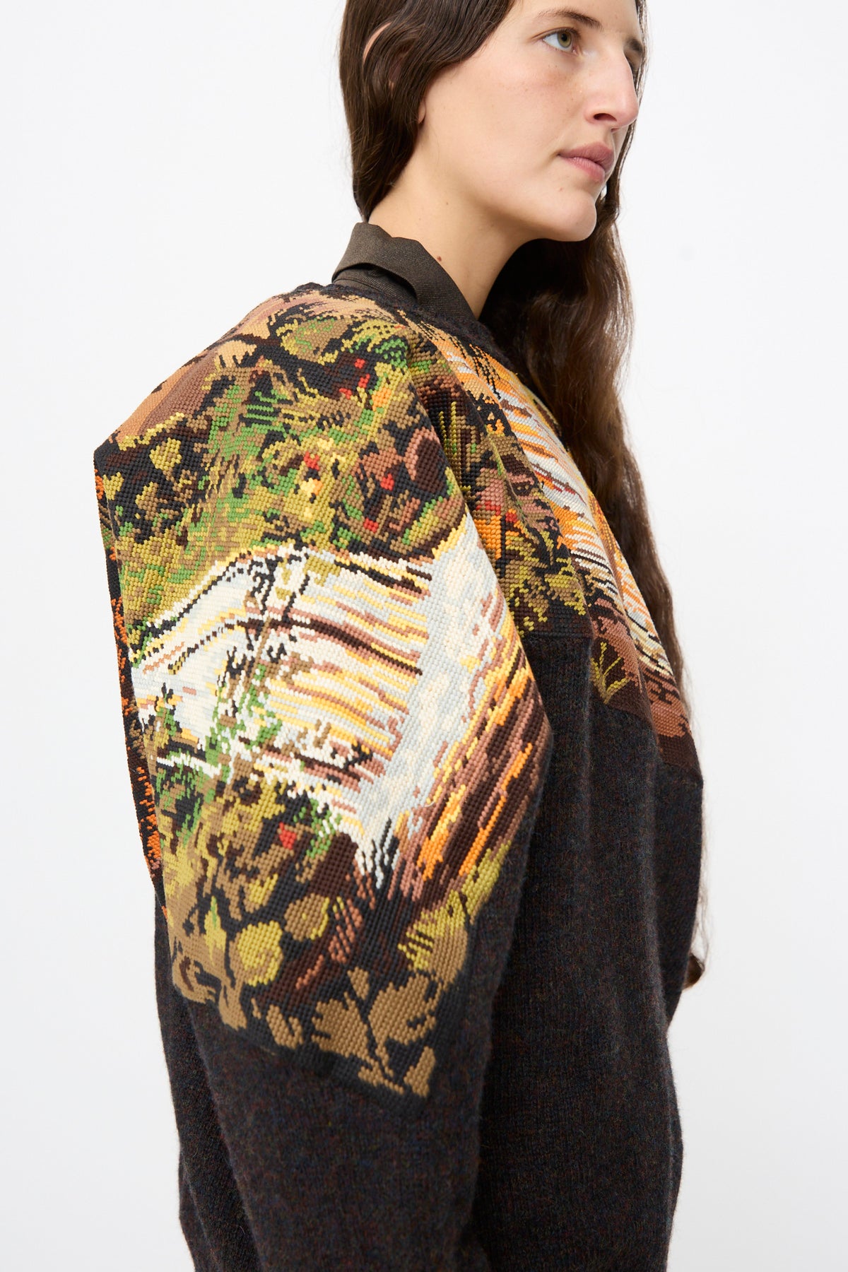 A person wearing a Bless No. 82 Kitschenrich in Unique Ultralarge Patchwork II dark wool pullover sweater with a colorful needlepoint landscape motif on the shoulder and sleeve.