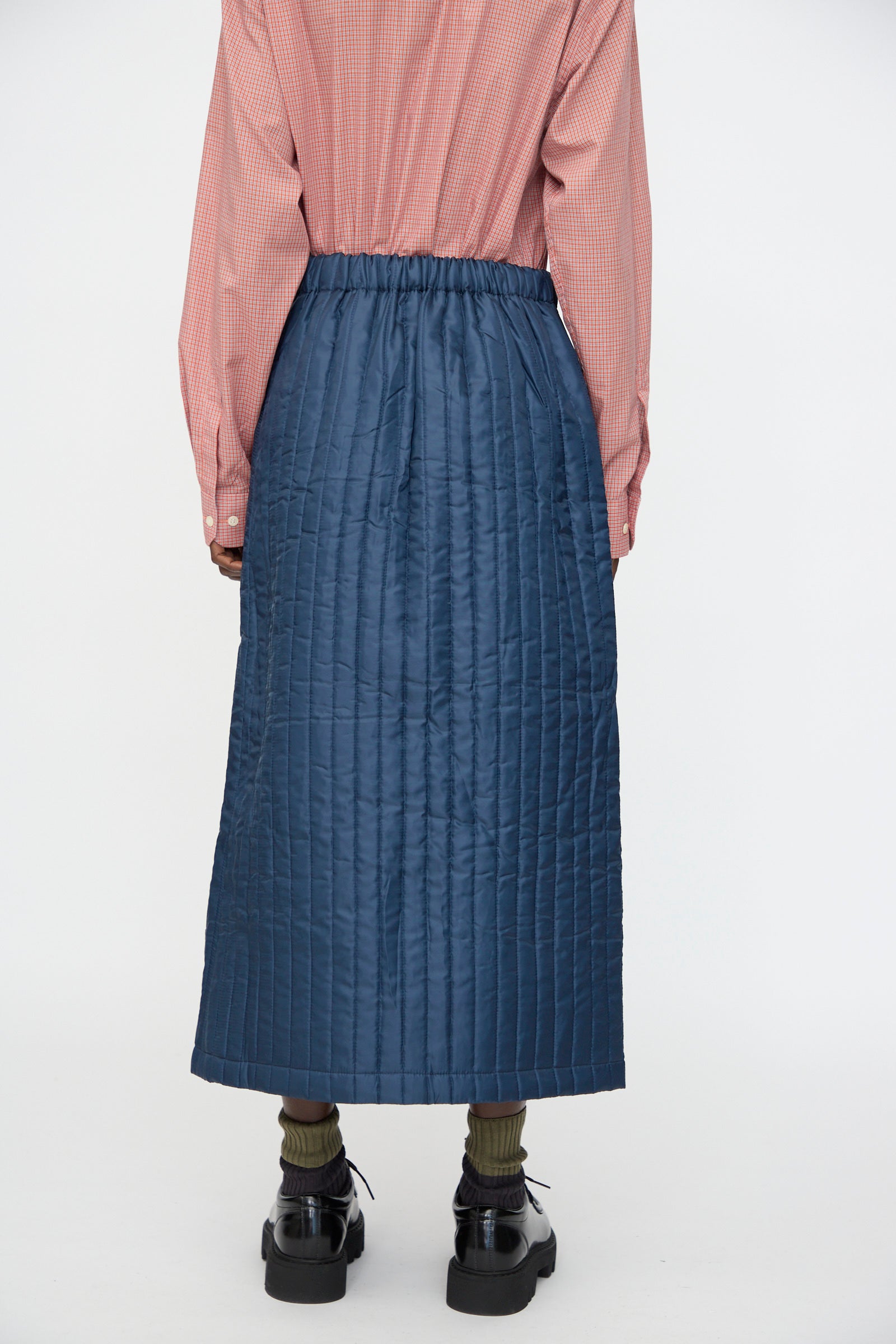 A person wears the Bless No. 08 Monpe Skirt in Navy with an adjustable snap waist, a pink checkered shirt, olive socks, and black shoes, standing with their back to a plain white background.