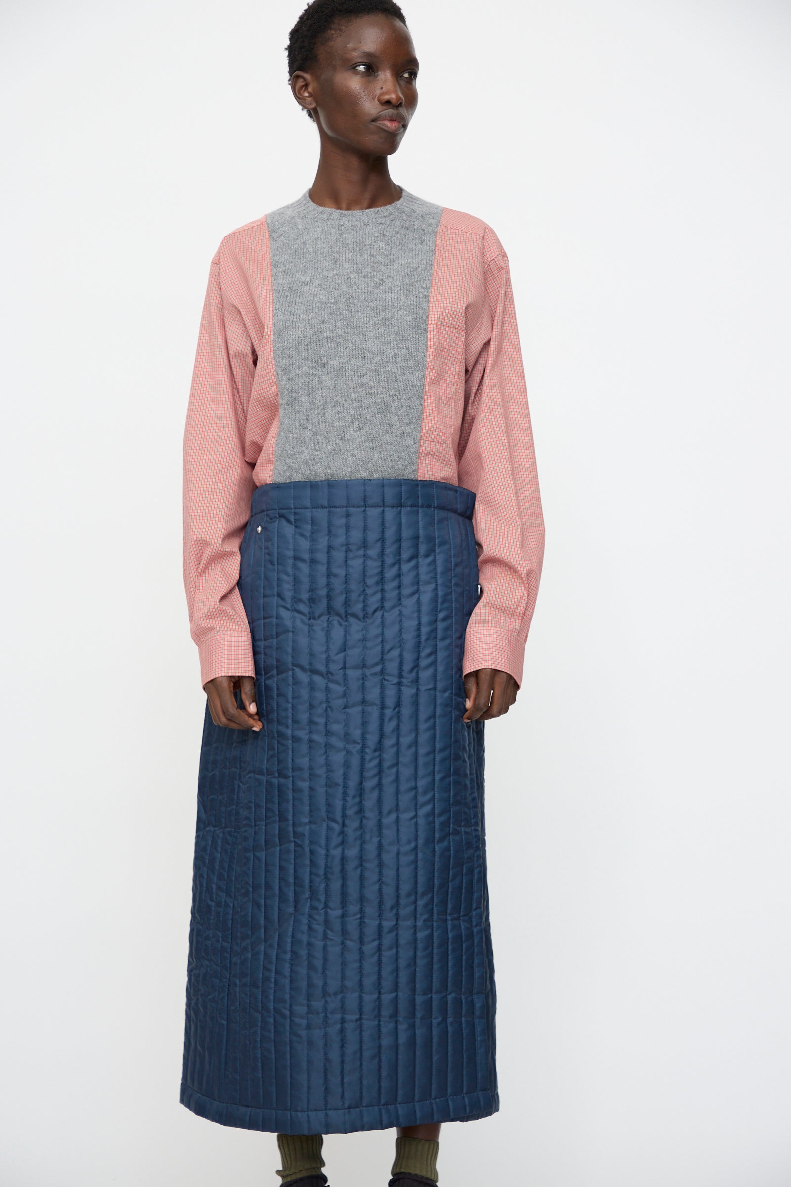 A person wears the Bless No. 08 Monpe Skirt in Navy, a blue quilted nylon skirt with an adjustable snap waist, paired with a grey and pink long-sleeve top, standing against a plain white background.