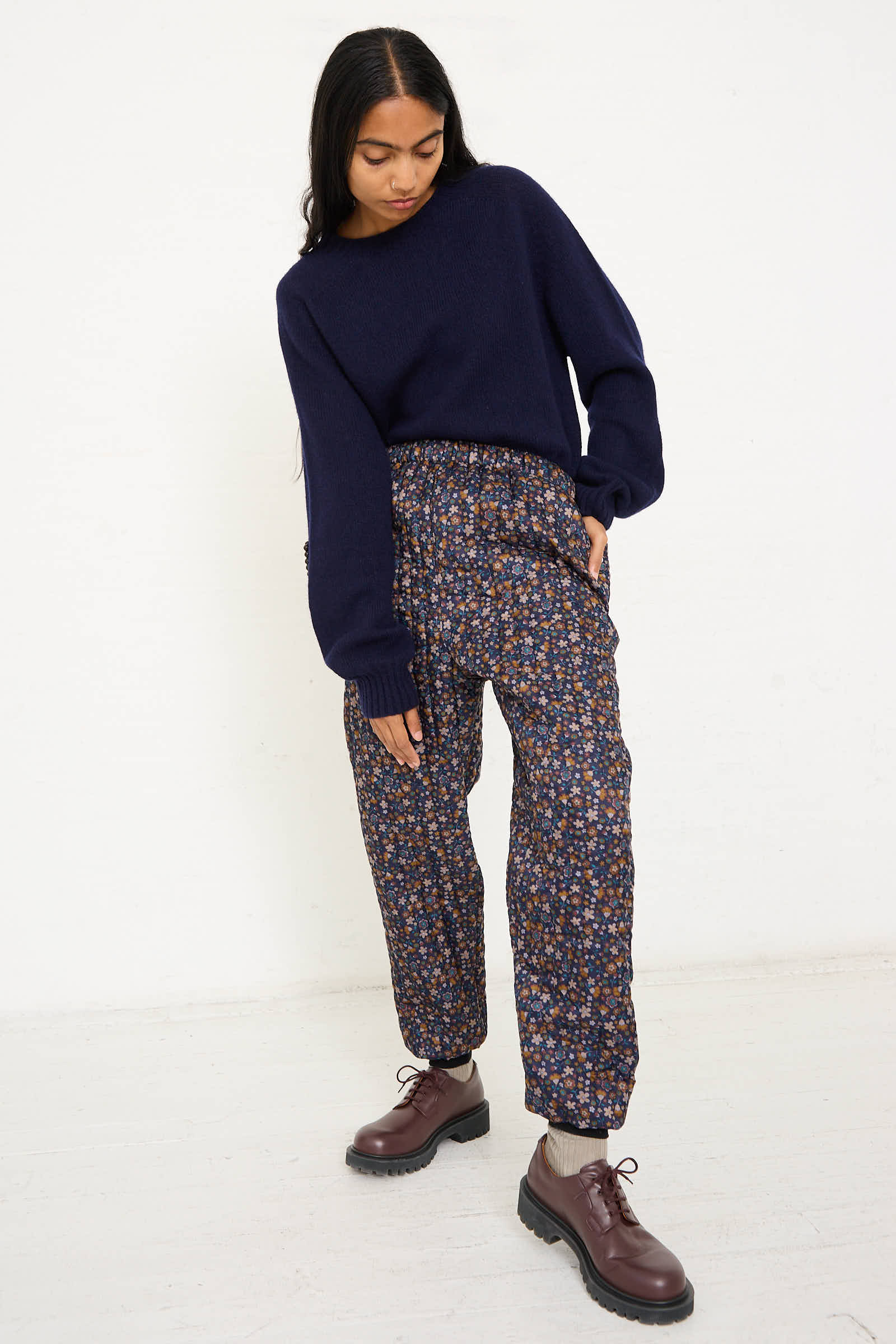 A person with long dark hair wears a navy sweater, No. 08 Monpe Pants in Navy Floral Multi by Bless, and brown shoes while standing in a white room.