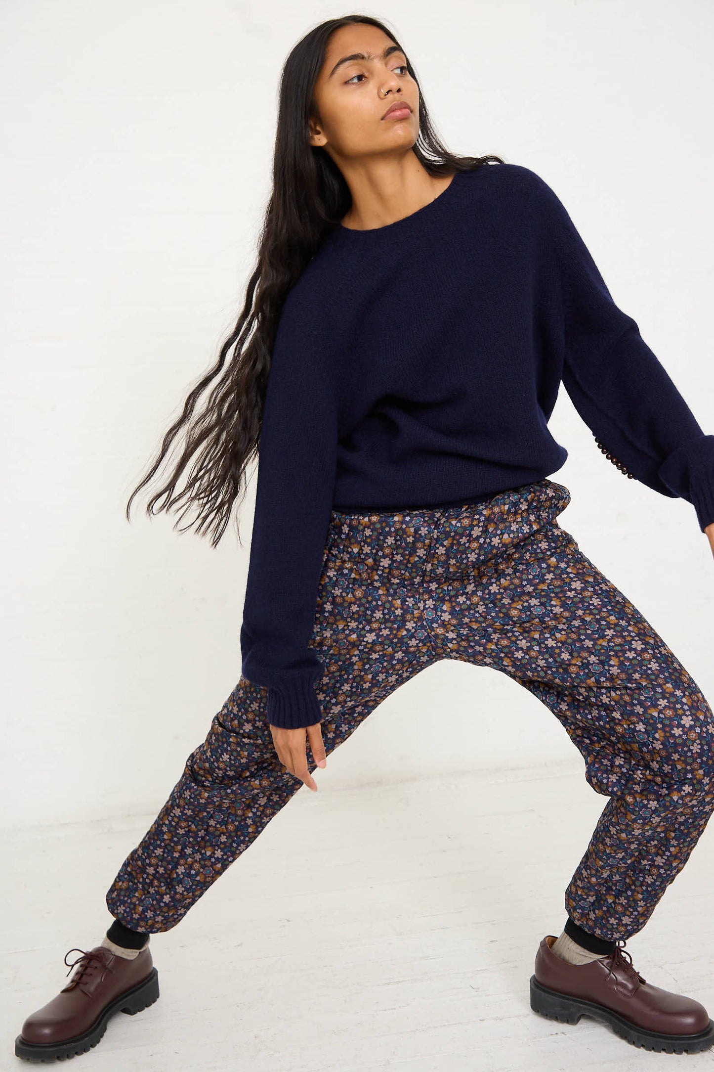 A person with long hair, wearing a dark sweater and the No. 08 Monpe Pant in Navy Floral Multi by Bless, along with brown shoes, posed dynamically against a plain background.