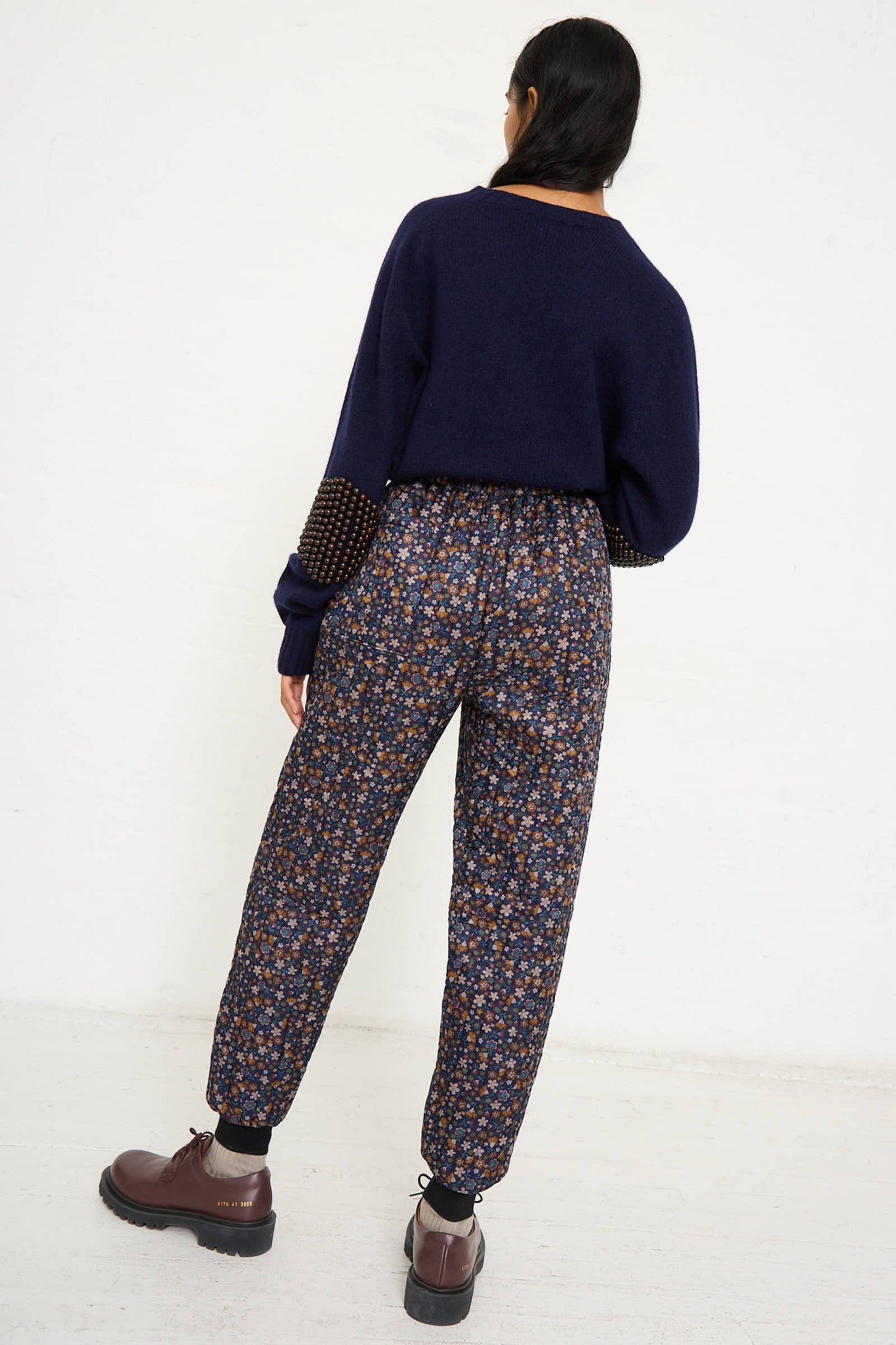 A person stands facing away wearing a navy sweater, No. 08 Monpe Pant in Navy Floral Multi by Bless, and brown shoes against a plain light-colored background.