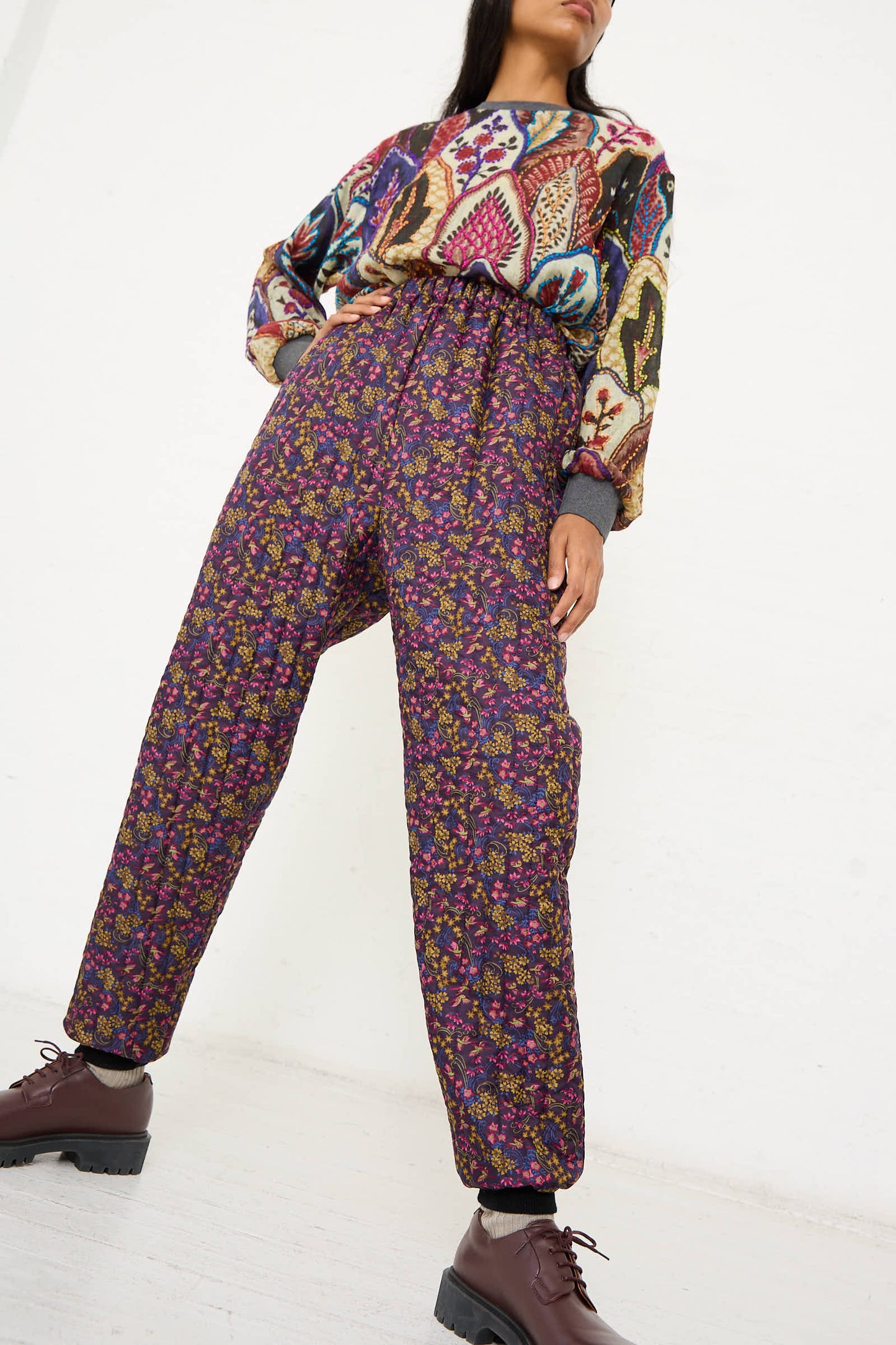 Person wearing a colorful long-sleeve top and the No. 08 Monpe Pant in Purple Floral Multi from Bless, with burgundy shoes, posed against a plain white background.