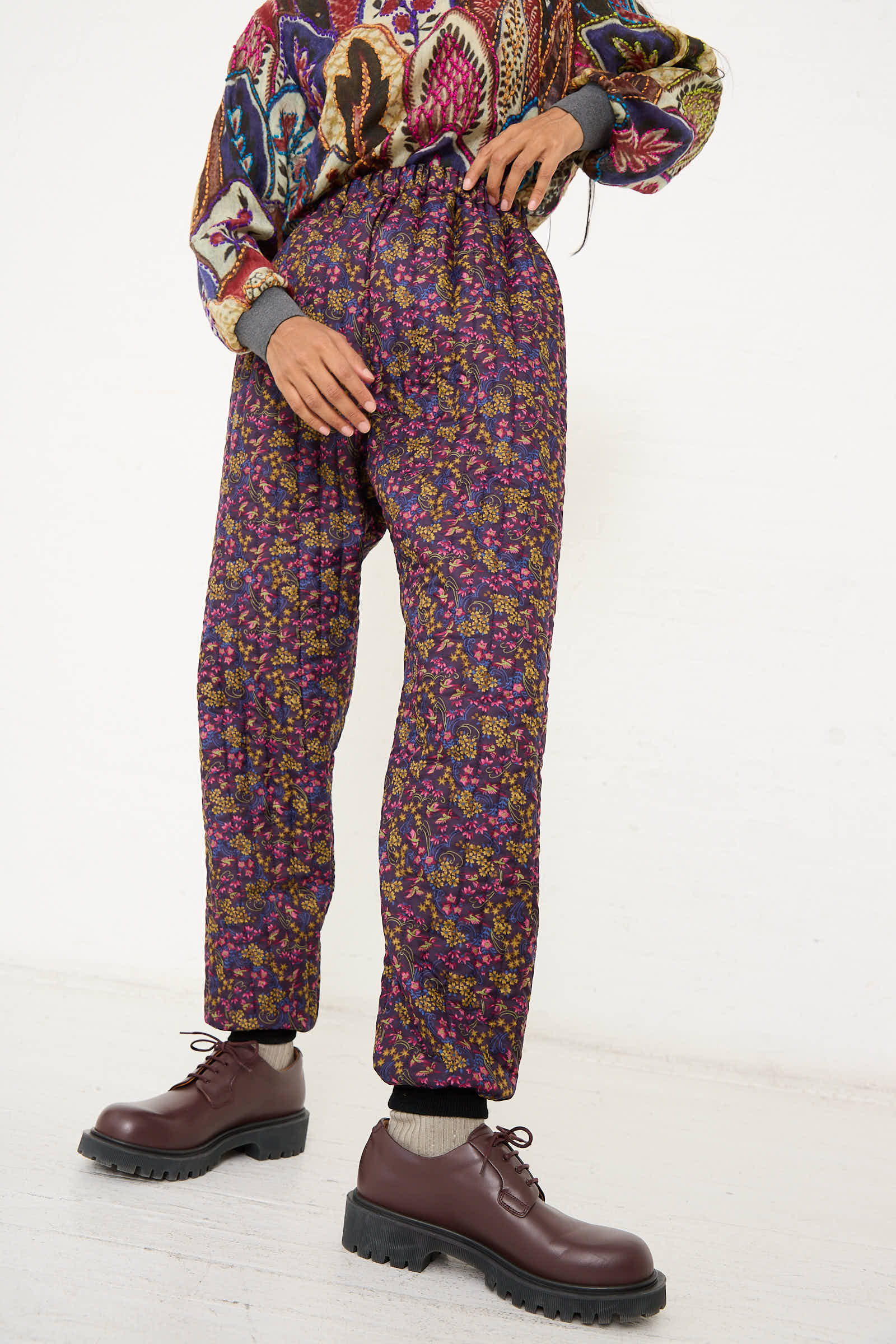 Person wearing a colorful sweater, gray socks, brown lace-up shoes, and the No. 08 Monpe Pant in Purple Floral Multi by Bless with an elasticized waist, standing against a plain white background.