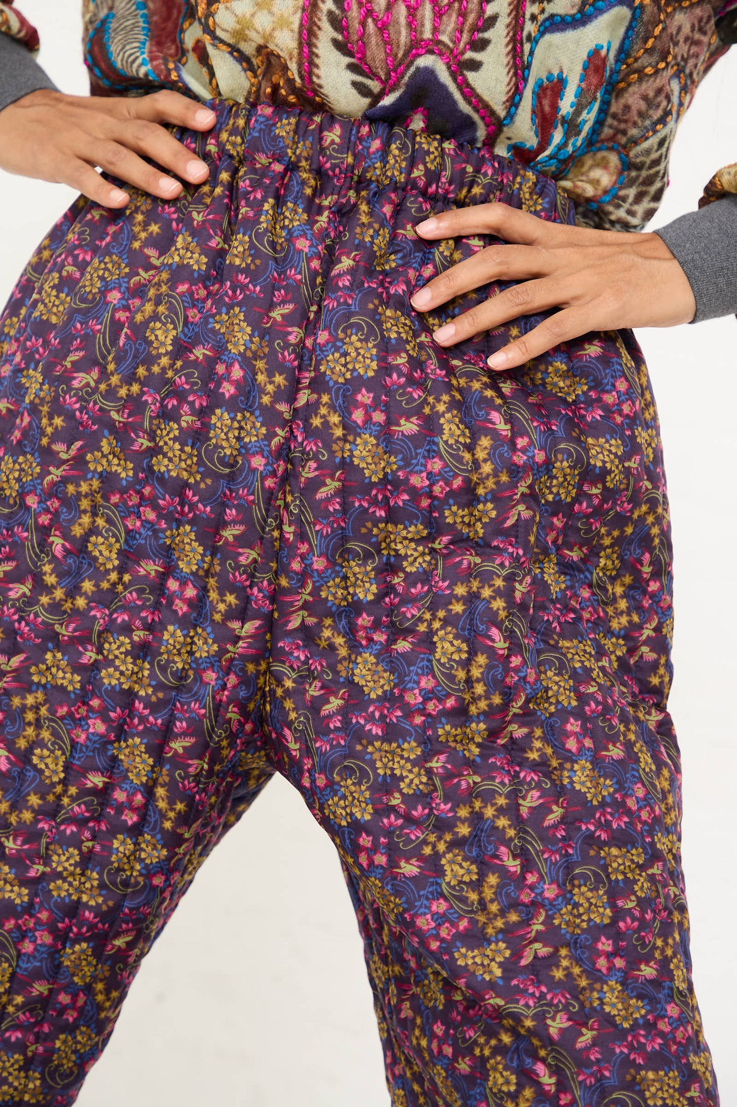 Close-up of a person wearing Bless's No. 08 Monpe Pant in Purple Floral Multi, with hands placed on hips. The pants, inspired by Japanese quilted farming pants, feature an elasticized waist and showcase vibrant purple floral patterns accented with shades of yellow and blue.