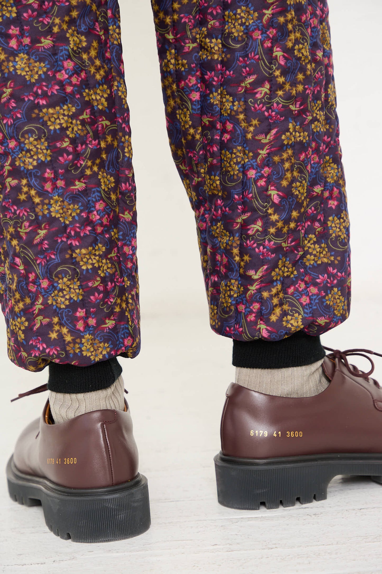 A person wearing the No. 08 Monpe Pant in Purple Floral Multi by Bless, with an elasticized waist, gray socks, and brown shoes with black soles.