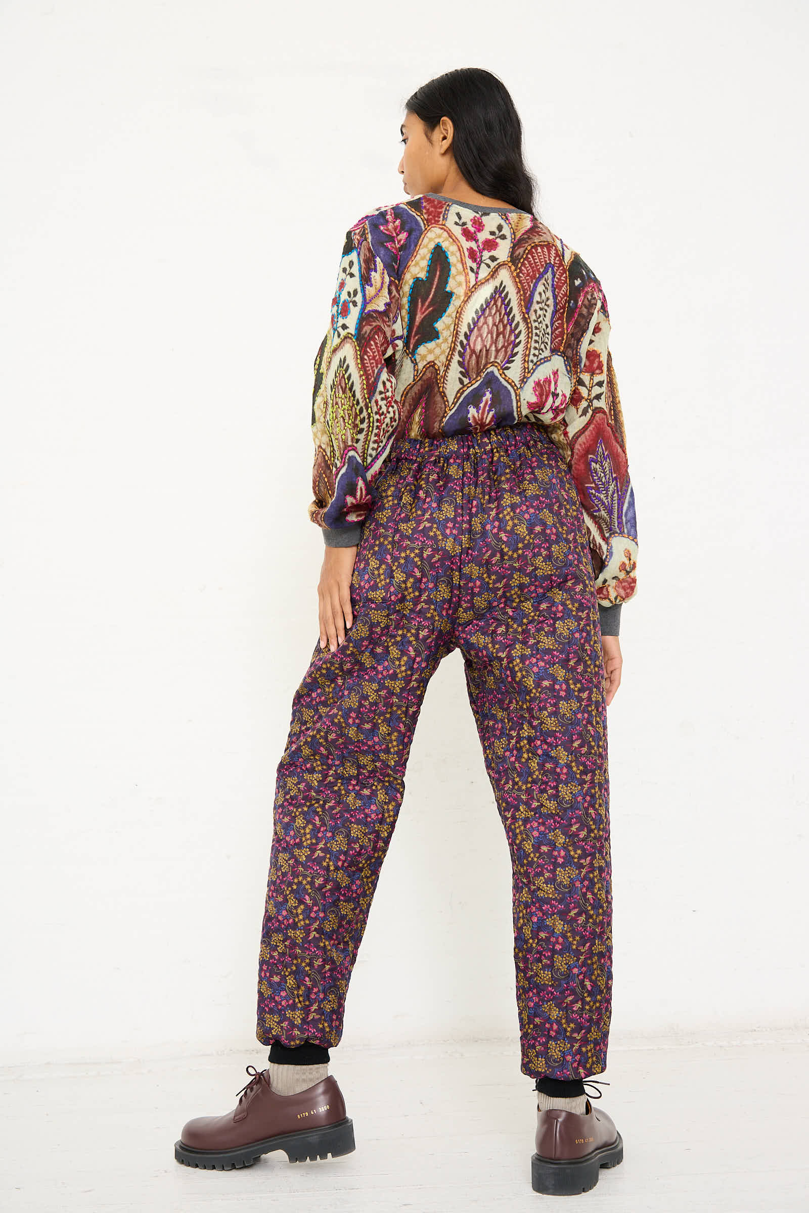 A person stands facing away from the camera, wearing a colorful floral-patterned sweater paired with Bless's No. 08 Monpe Pant in Purple Floral Multi, featuring an elasticized waist, and finished off with brown shoes.