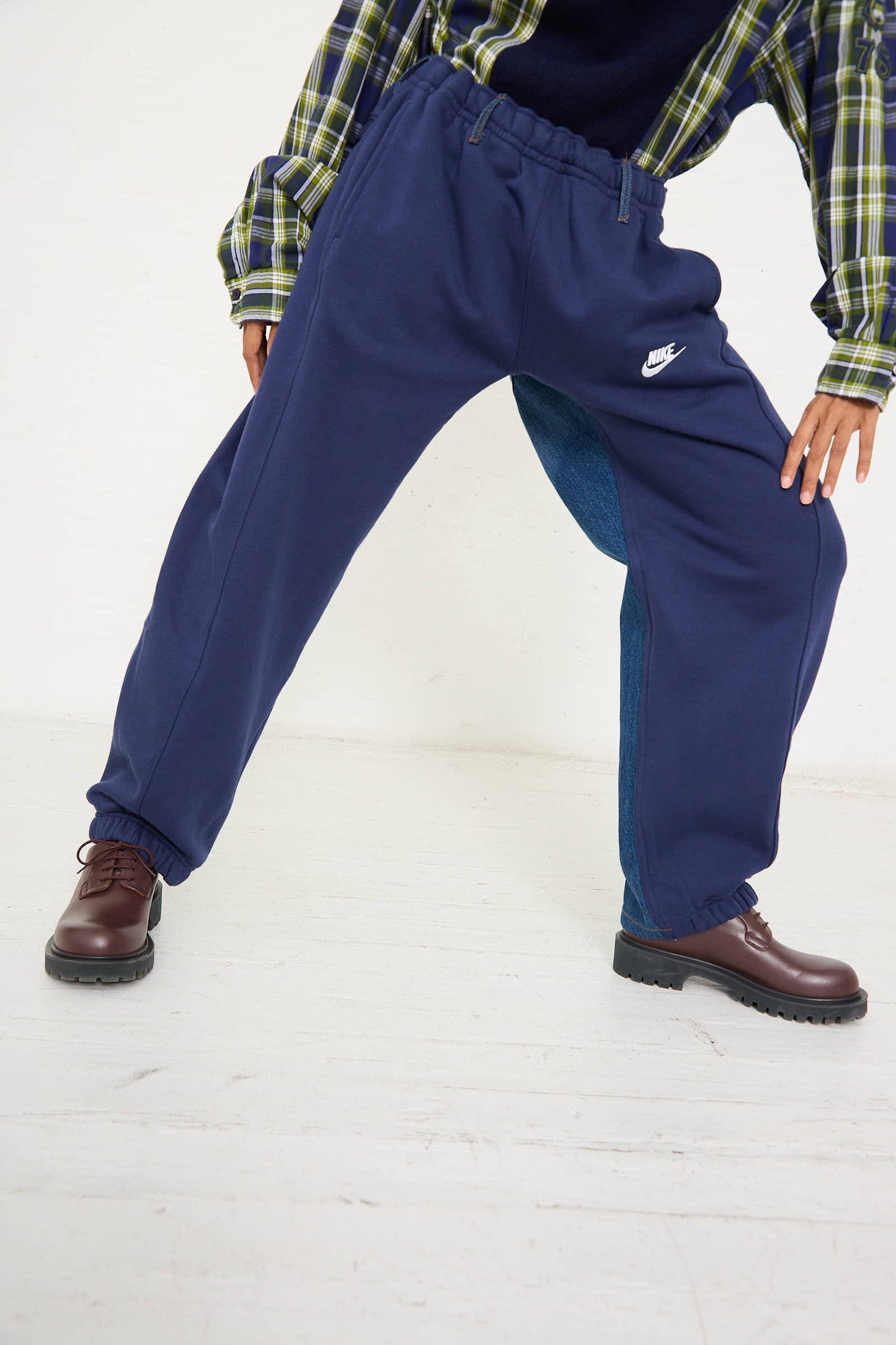 Person wearing No. 68 Overjoggingjeans in Blue and Black by Bless, a green plaid shirt, and brown shoes stands with legs spread apart, leaning slightly forward, against a plain background.