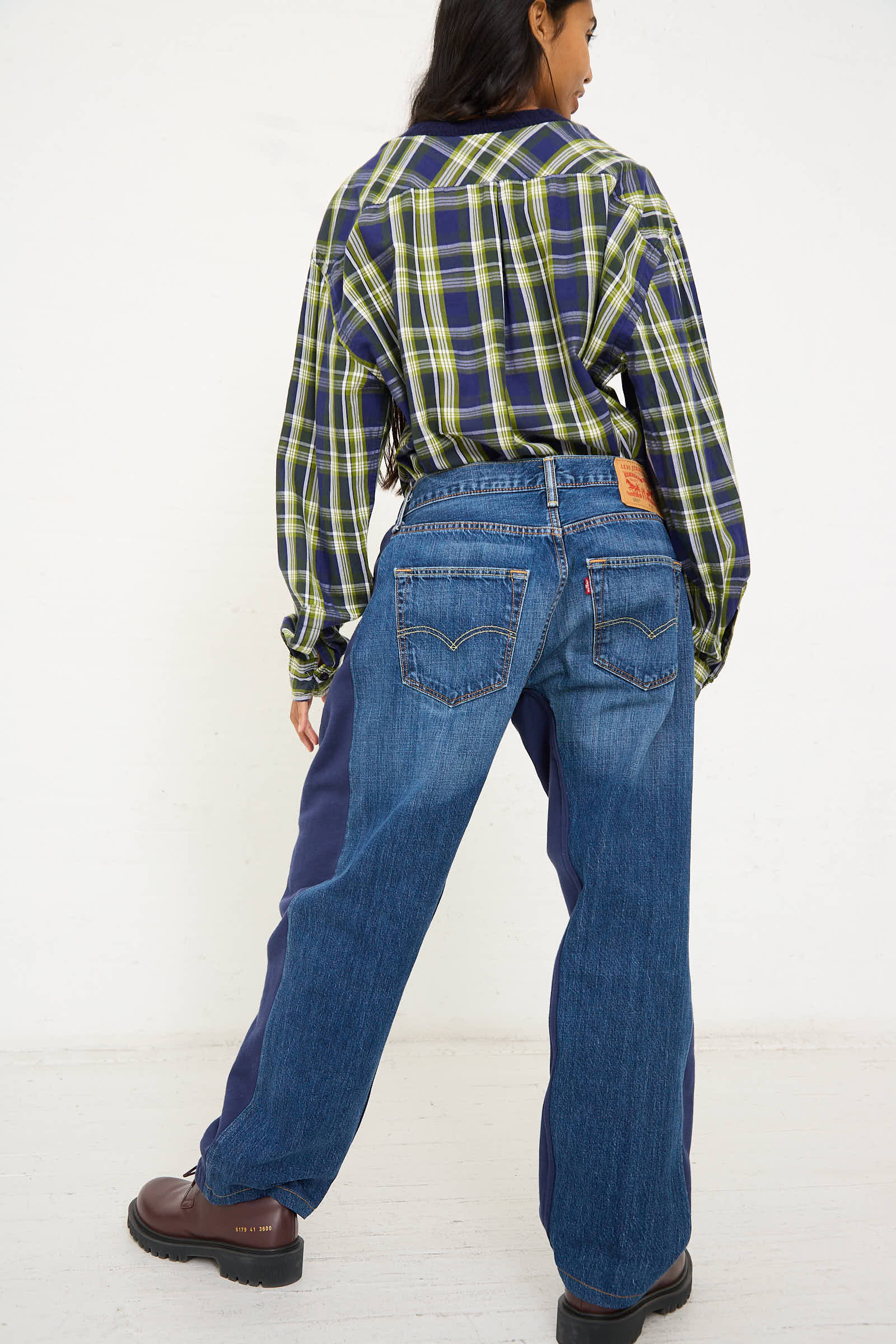 A person wearing a plaid shirt, Bless's No. 68 Overjoggingjeans in Blue and Black with visible back pockets, and brown shoes is standing with their back to the camera.